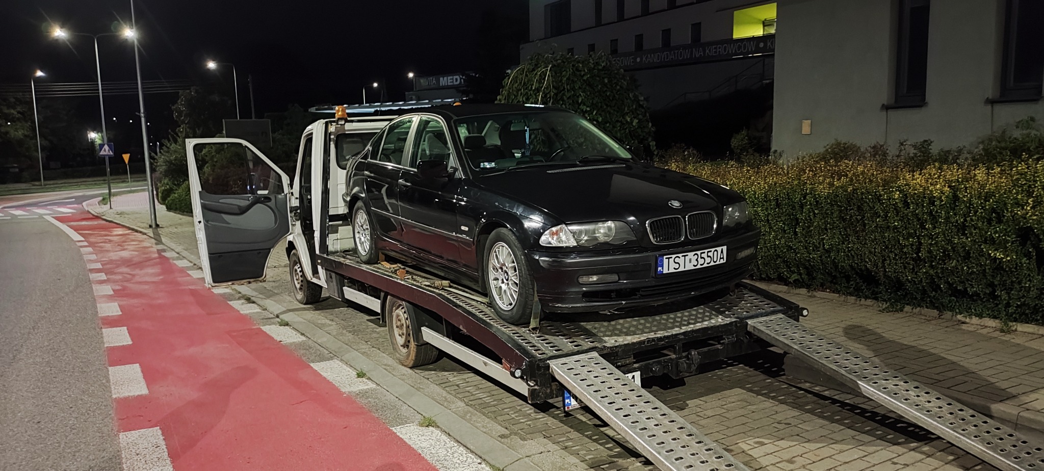 Czarny samochód BMW na lawecie w nocy, z otwartymi drzwiami busa. Widoczny chodnik z czerwoną ścieżką rowerową i budynek w tle.