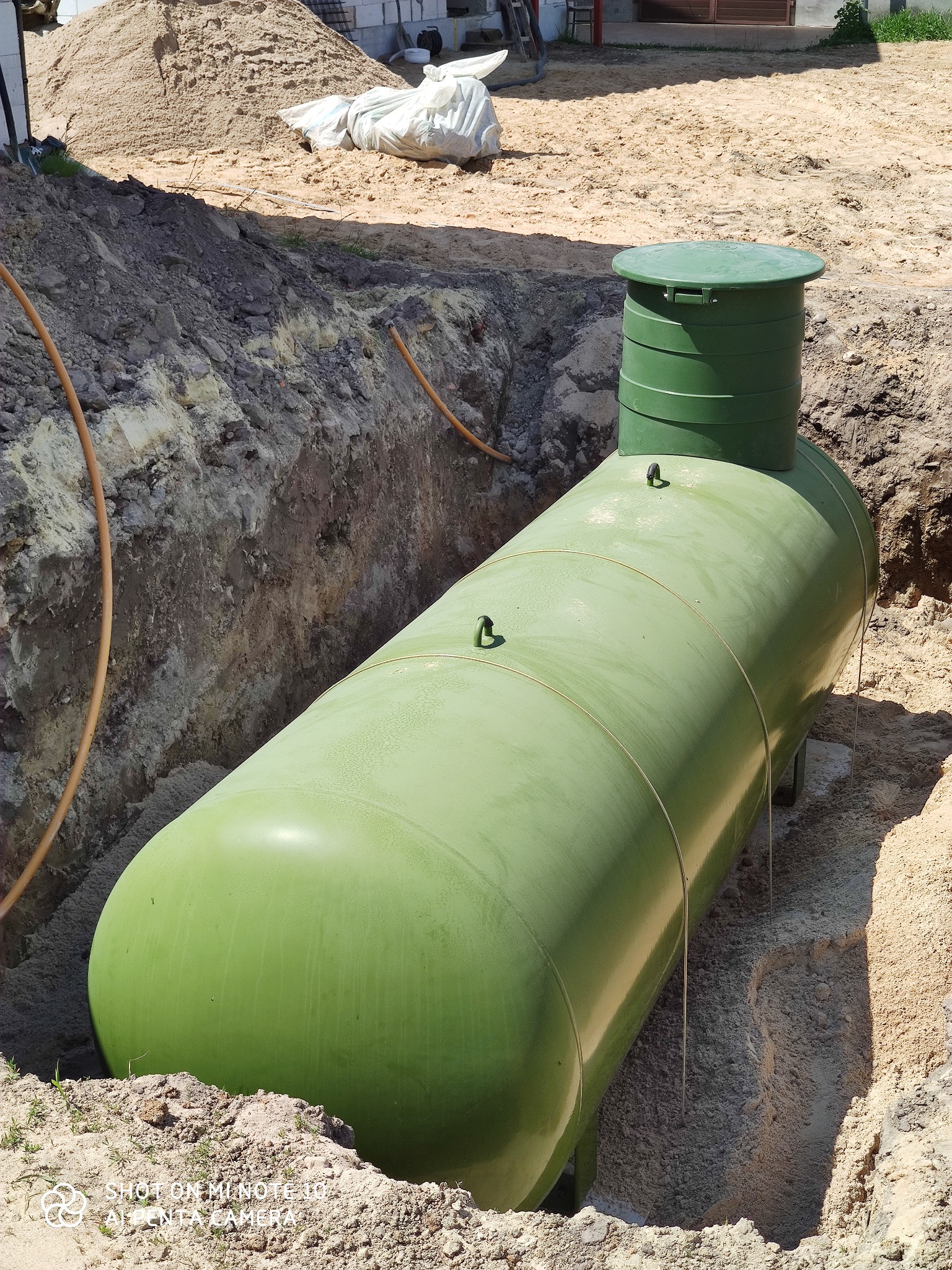 Zielony zbiornik zakopany w wykopie z piasku, widoczny fragment rury. Zbiornik posiada zieloną pokrywę. W tle widać elementy konstrukcji budynku.