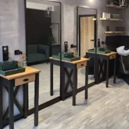 Stylowe stanowiska fryzjerskie z dużymi lustrami w czarnych ramach, drewnianymi blatami i czarnymi nogami w kształcie litery X. Widoczne półki z kosmetykami i fotel do mycia włosów.