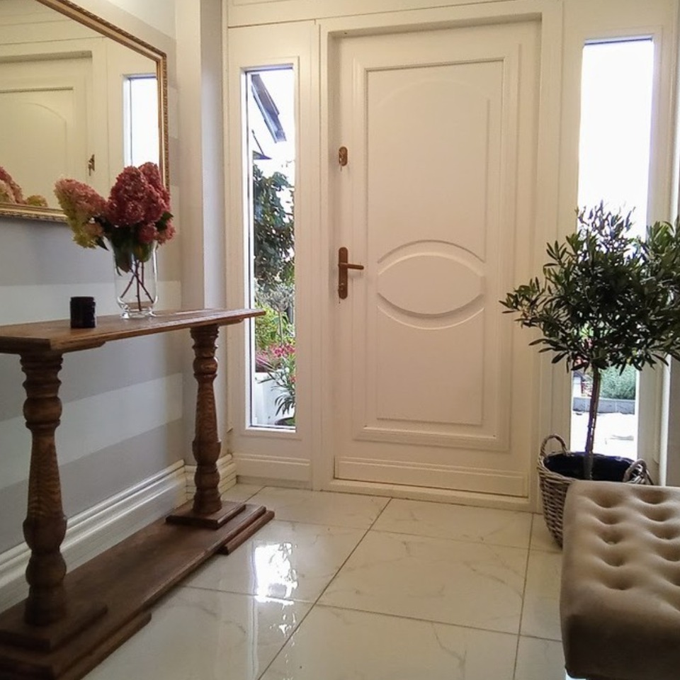 Białe drzwi wejściowe z szybkami bocznymi, drewniana konsola z kwiatami, lustro w złotej ramie i marmurowa podłoga tworzą elegancki wystrój przedpokoju.