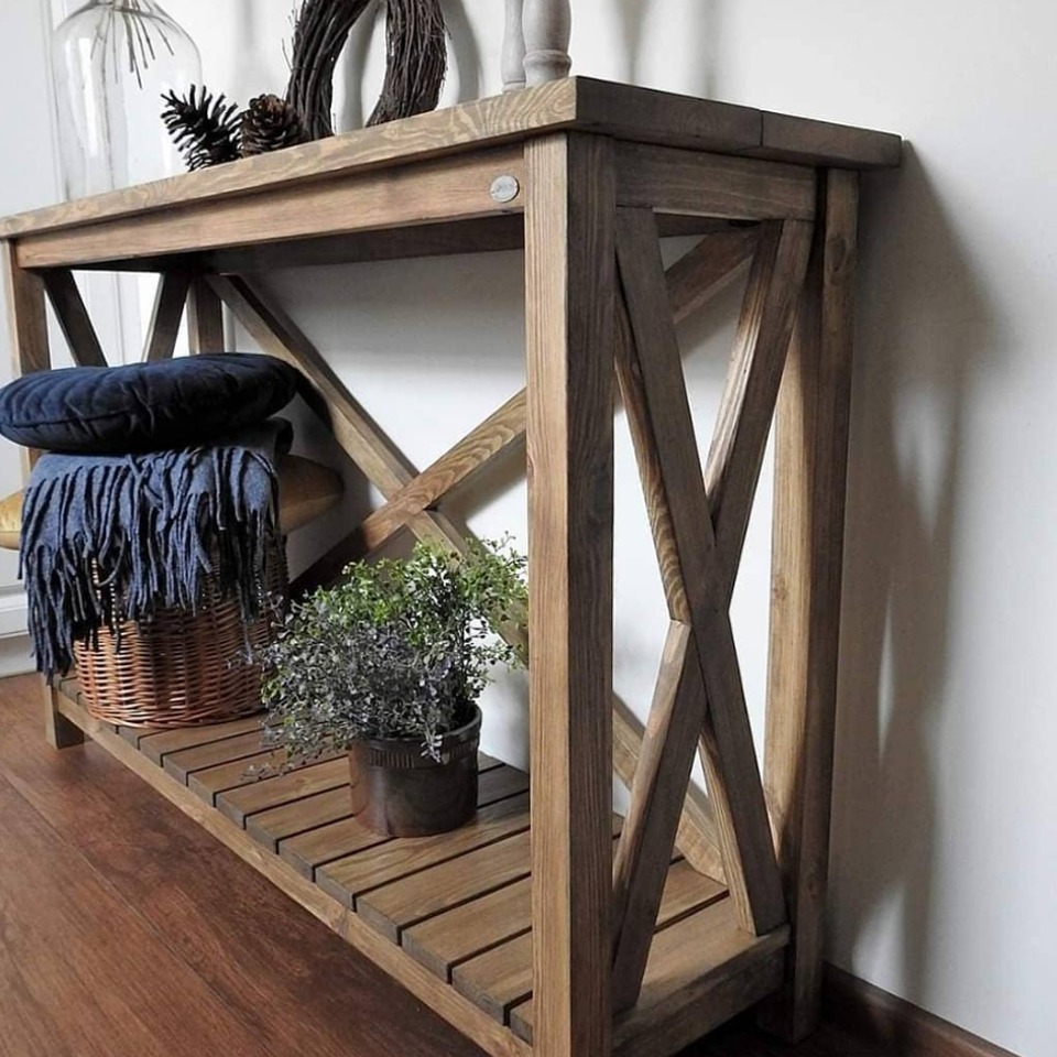 Rustykalny stolik konsola z drewna o ciepłym odcieniu, z dekoracjami: poduszką, pledem i rośliną doniczkową. Wyeksponowane detale konstrukcyjne w kształcie litery X.