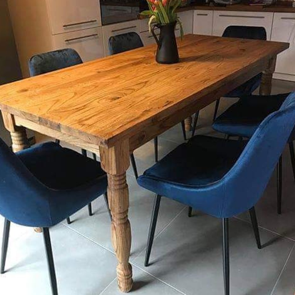 Rustykalny stół z litego drewna z toczonymi nogami w jadalni, otoczony niebieskimi krzesłami na tle białej zabudowy kuchennej. Na stole czarny wazon z tulipanami.