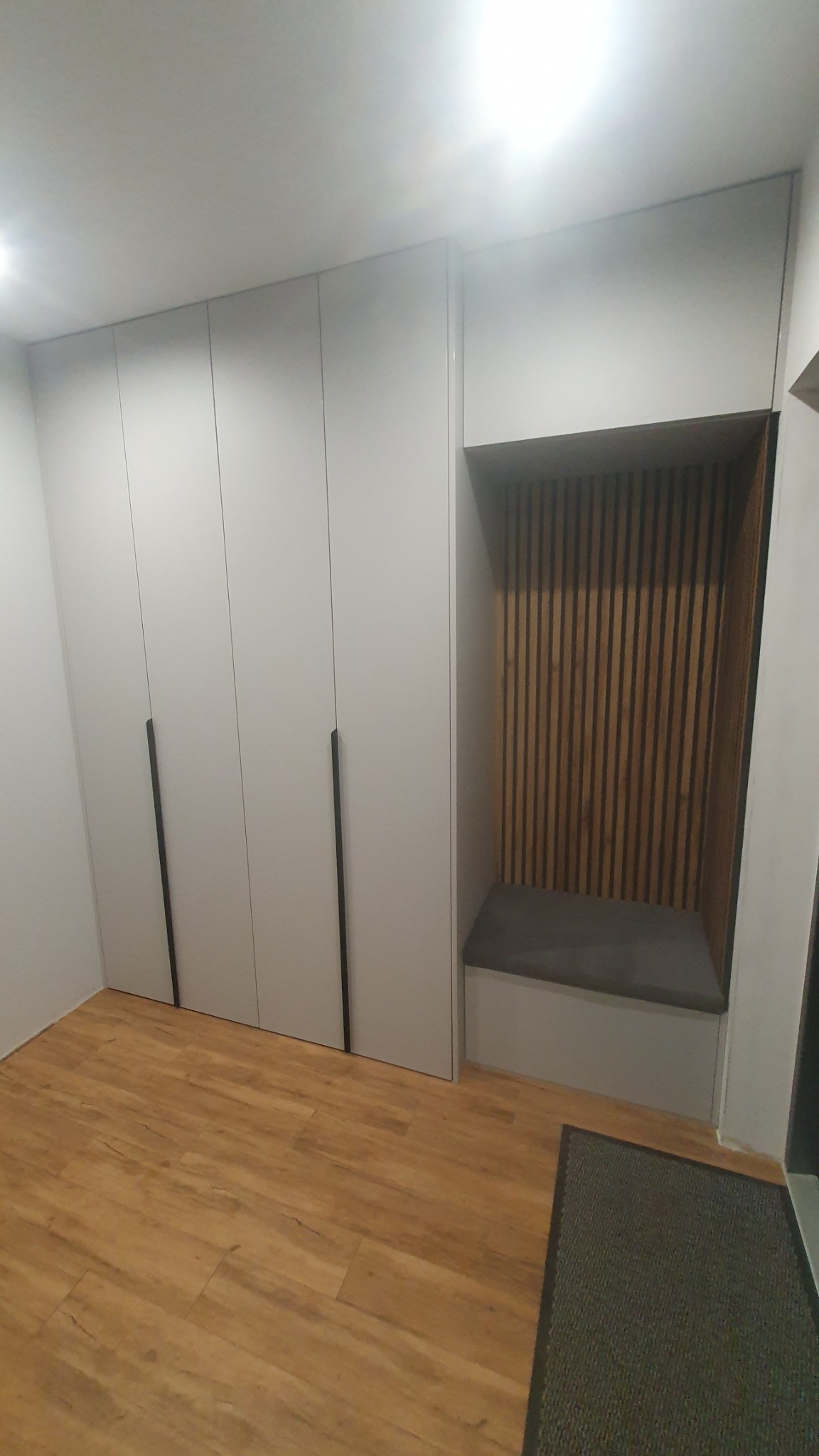 Biała szafa wnękowa z siedziskiem i drewnianymi lamelami w przedpokoju, widok na podłogę z paneli. Minimalistyczny design, czarne uchwyty.