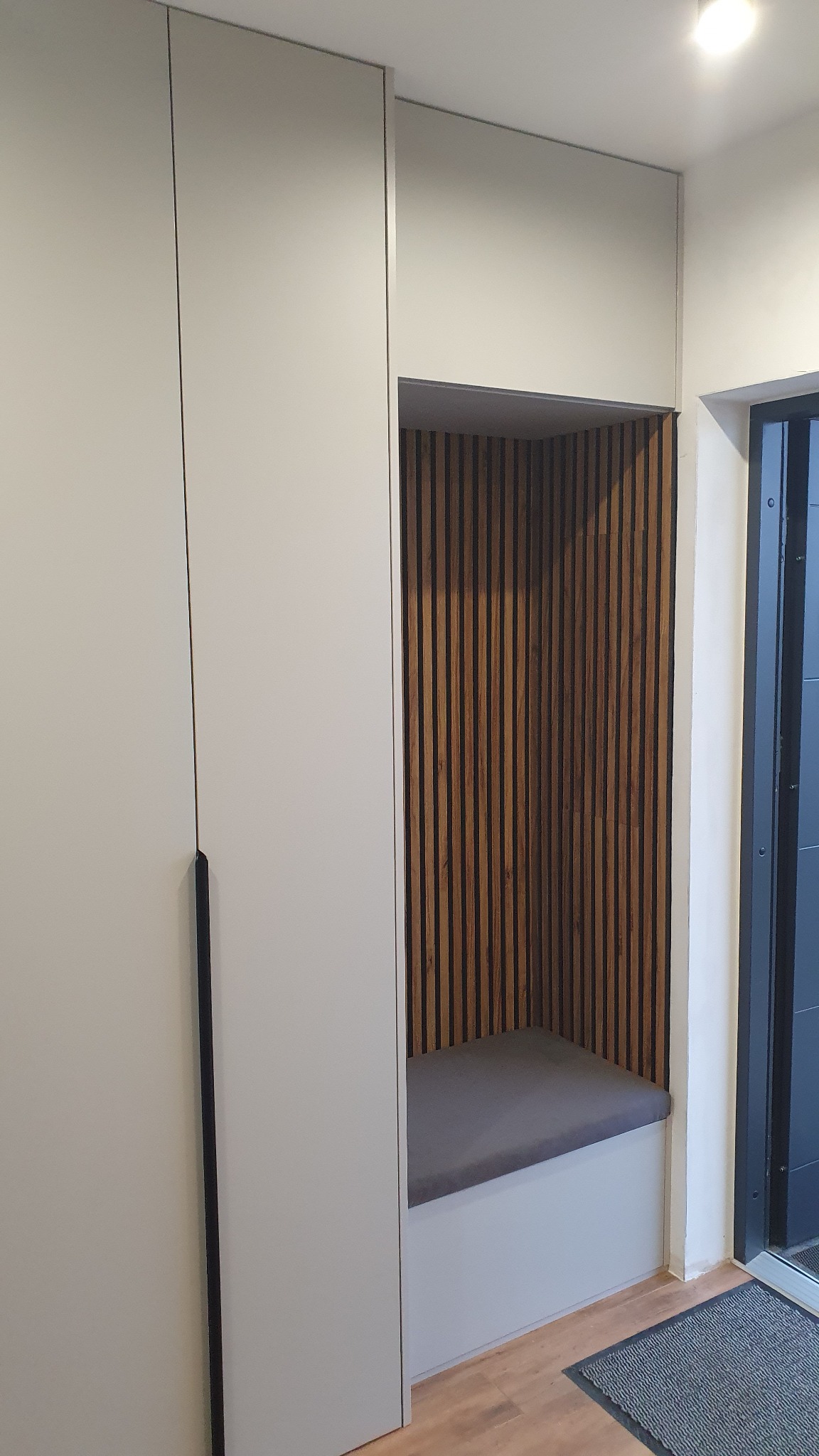 Jasna szafa wnękowa z siedziskiem i drewnianymi lamelami na ścianie wnęki, minimalistyczny design, widok z bliska, szary dywanik przy drzwiach.