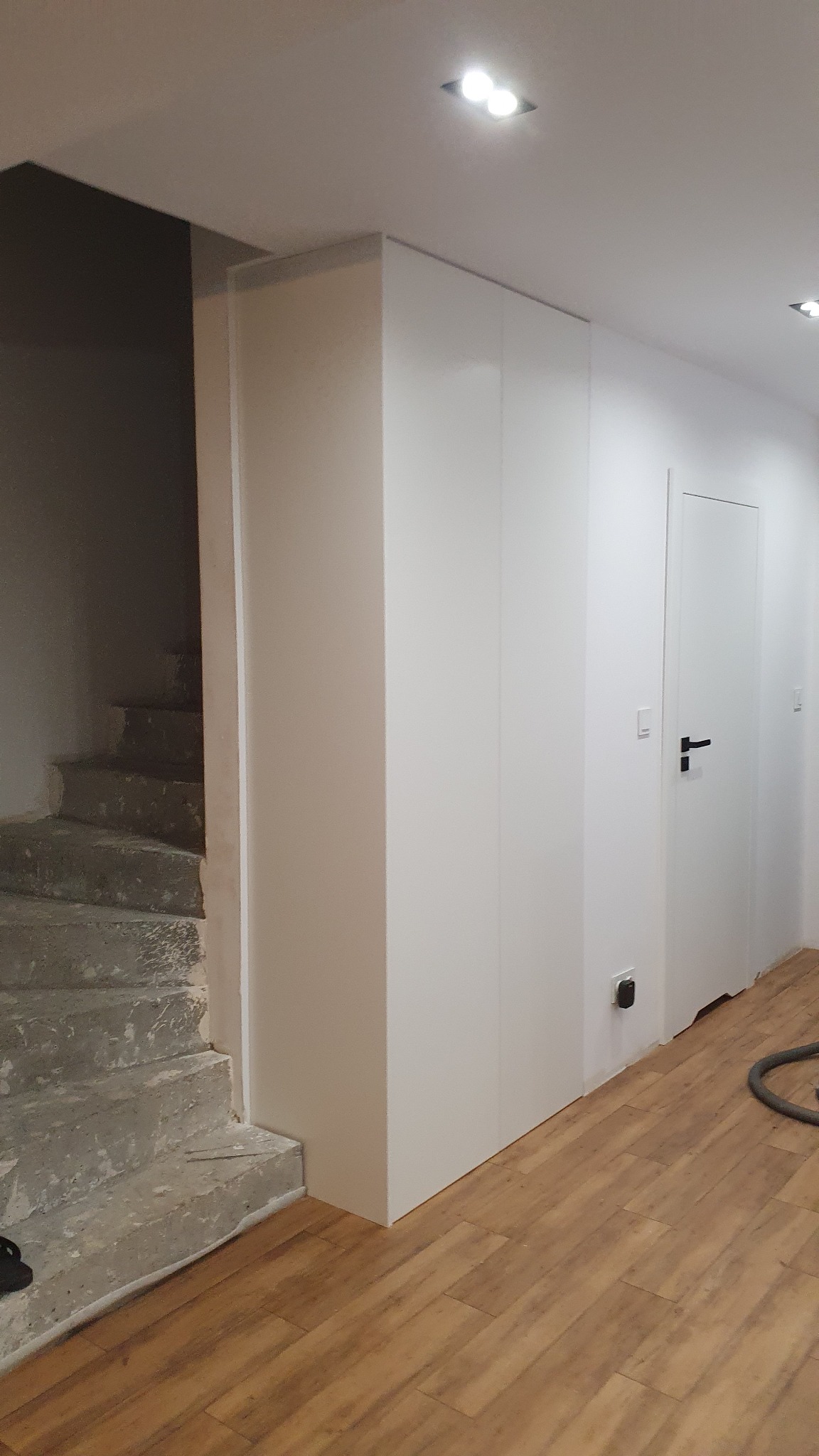 Biała szafa wnękowa z drzwiami, wkomponowana w korytarz z drewnianą podłogą i betonowymi schodami. Minimalistyczny design, oświetlenie sufitowe.