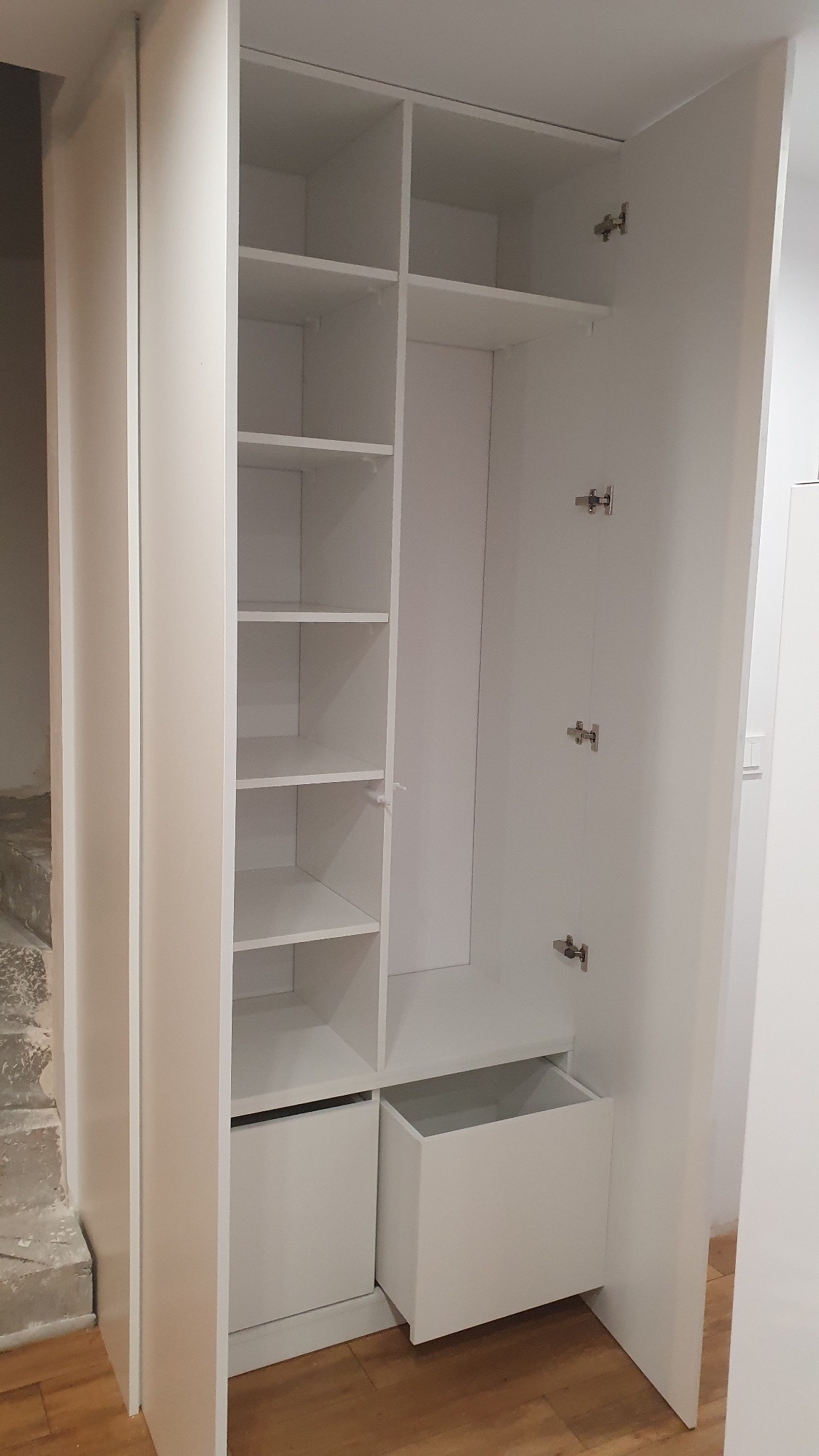 Biała szafa wnękowa z otwartymi drzwiami, widoczne półki i szuflady. Minimalistyczny design, jasne wnętrze, metalowe zawiasy. Częściowo widoczna podłoga.