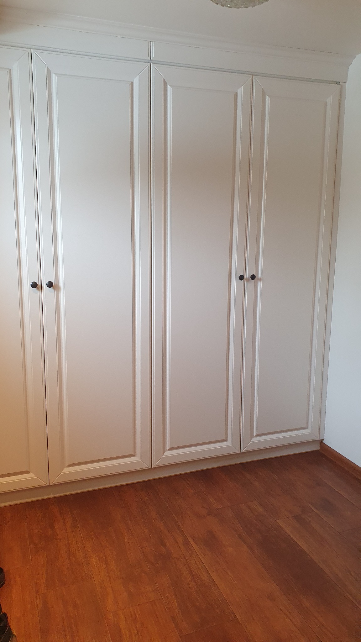 Biała szafa wnękowa z czterema drzwiami, z ciemnymi okrągłymi uchwytami, na tle drewnianej podłogi i białej ściany. Klasyczny styl.