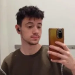 Mężczyzna robi selfie w lustrze, trzymając smartfon w złotej obudowie. Widoczne słuchawki bezprzewodowe w uchu, ciemne włosy i oliwkowa koszulka. Tło stanowi jasna ściana.