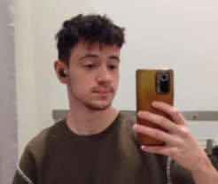 Mężczyzna robi selfie w lustrze, trzymając smartfon w złotej obudowie. Widoczne słuchawki bezprzewodowe w uchu, ciemne włosy i oliwkowa koszulka. Tło stanowi jasna ściana.