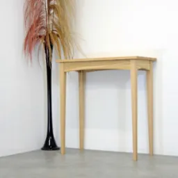 Konsola z jasnego drewna w minimalistycznym stylu, ustawiona przy białej ścianie obok wysokiego, czarnego wazonu z suszonymi trawami.