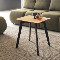 Kwadratowy stolik kawowy z blatem z jasnego drewna i ciemnymi nogami, na beżowym dywanie, z zieloną sofą w tle. Na blacie czarna miseczka i szklana ozdoba.