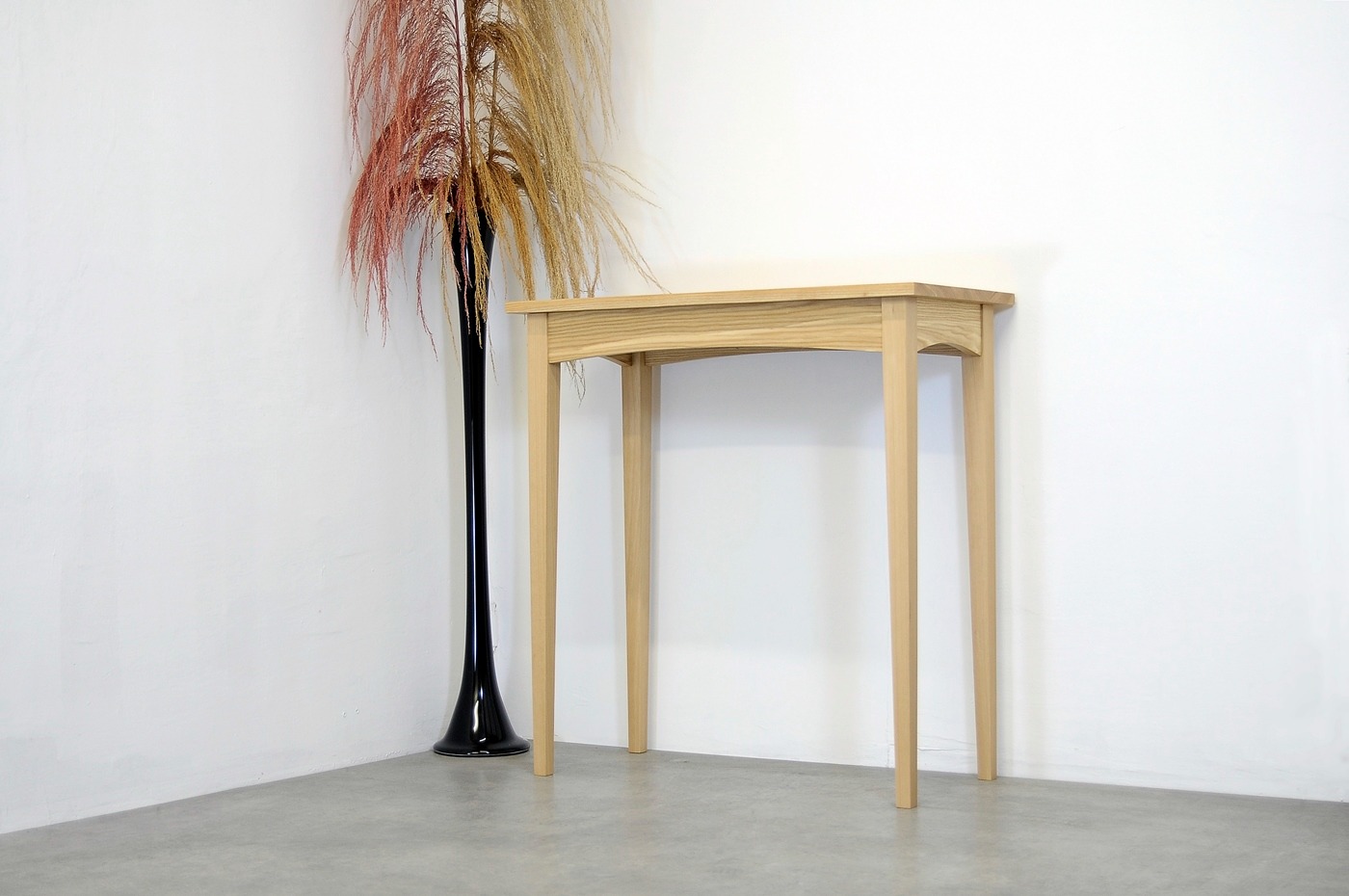 Konsola z jasnego drewna w minimalistycznym stylu, ustawiona przy białej ścianie obok wysokiego, czarnego wazonu z suszonymi trawami.