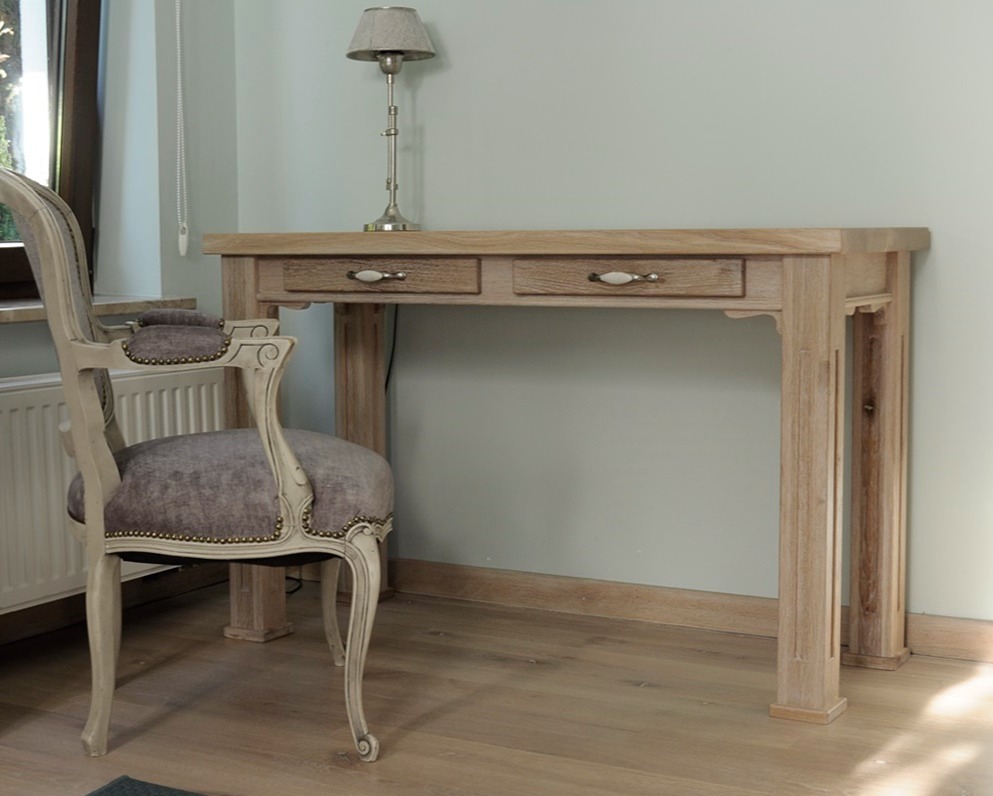 Jasne, drewniane biurko z dwiema szufladami w stylu retro, z ozdobnym krzesłem i lampką na blacie, na tle jasnej ściany i podłogi z paneli.