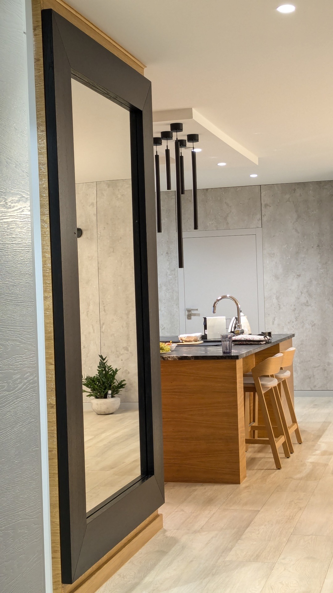 Nowoczesna kuchnia z drewnianą wyspą i hokerami, widziana przez czarną ramę drzwiową. Minimalistyczne oświetlenie i rośliny doniczkowe dodają elegancji.