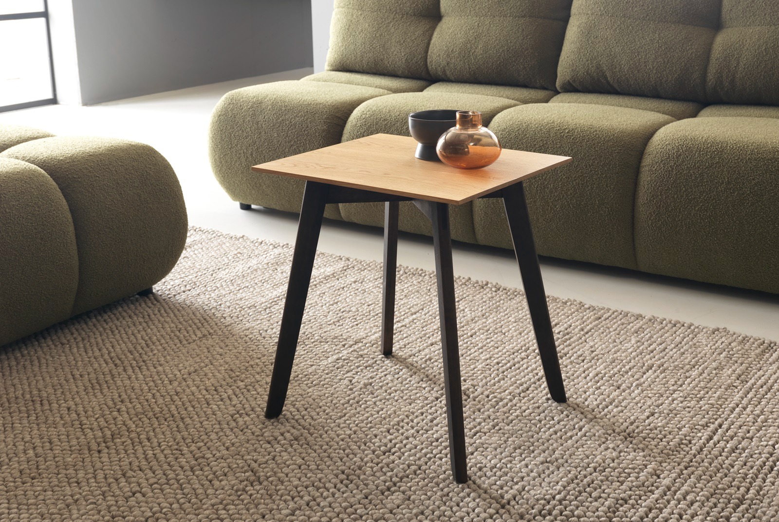 Kwadratowy stolik kawowy z blatem z jasnego drewna i ciemnymi nogami, na beżowym dywanie, z zieloną sofą w tle. Na blacie czarna miseczka i szklana ozdoba.