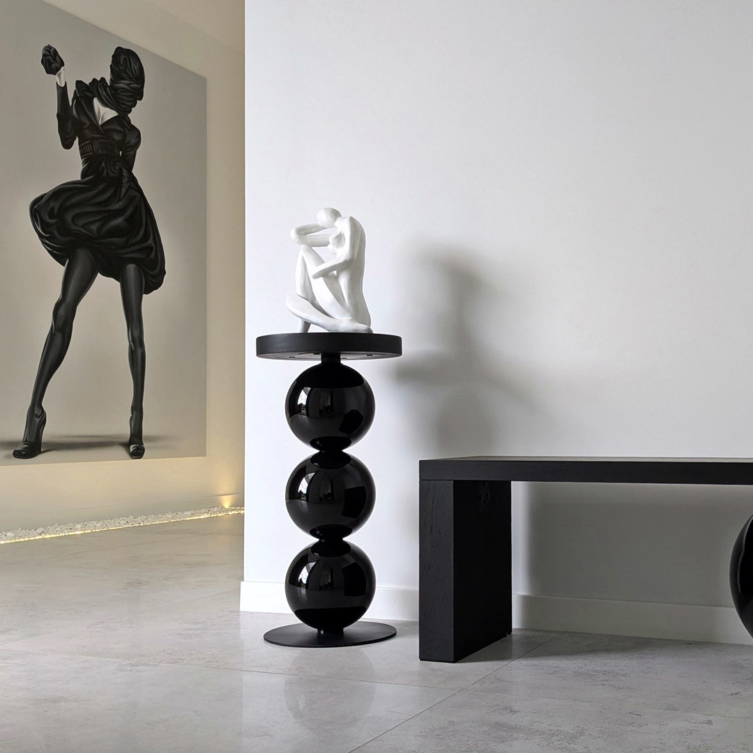 Nowoczesna aranżacja wnętrza: czarny stolik z kulami, biała rzeźba i obraz z sylwetką kobiety w czarnej sukni. Minimalistyczny design, gładka podłoga, jasne ściany.