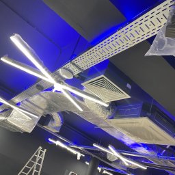 Michał Danowski - Rozbudowana instalacja oświetleniowa LED na suficie z widocznymi elementami wentylacji i drabiny, oświetlona niebieskim światłem, sugerująca prace instalacyjne w toku.