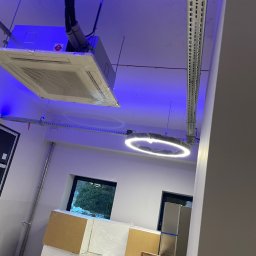 Michał Danowski - Kasetonowa klimatyzacja sufitowa w folii ochronnej, okrągła lampa LED. Widoczne kable i elementy montażowe. Niebieskie oświetlenie sufitowe.
