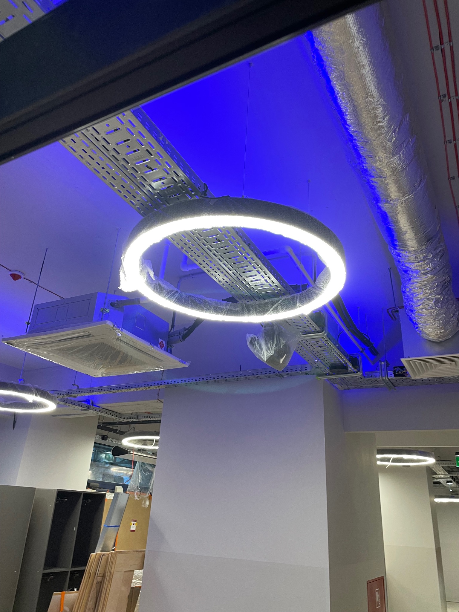 Okrągła lampa LED pod sufitem z instalacjami, w tle niebieskie oświetlenie. Widoczne koryta kablowe, fragment klimatyzacji w folii. Nowoczesne oświetlenie biurowe.