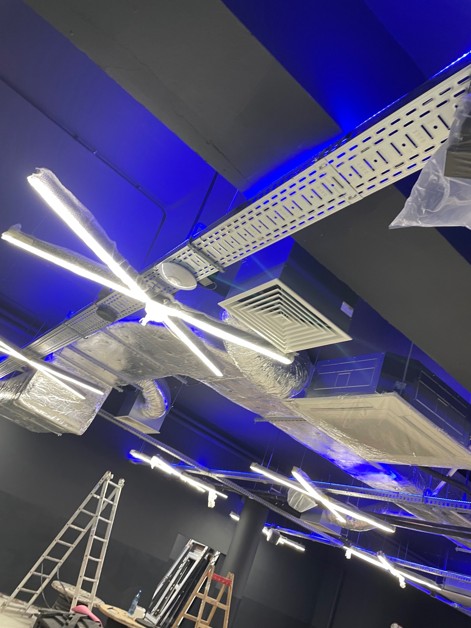 Rozbudowana instalacja oświetleniowa LED na suficie z widocznymi elementami wentylacji i drabiny, oświetlona niebieskim światłem, sugerująca prace instalacyjne w toku.