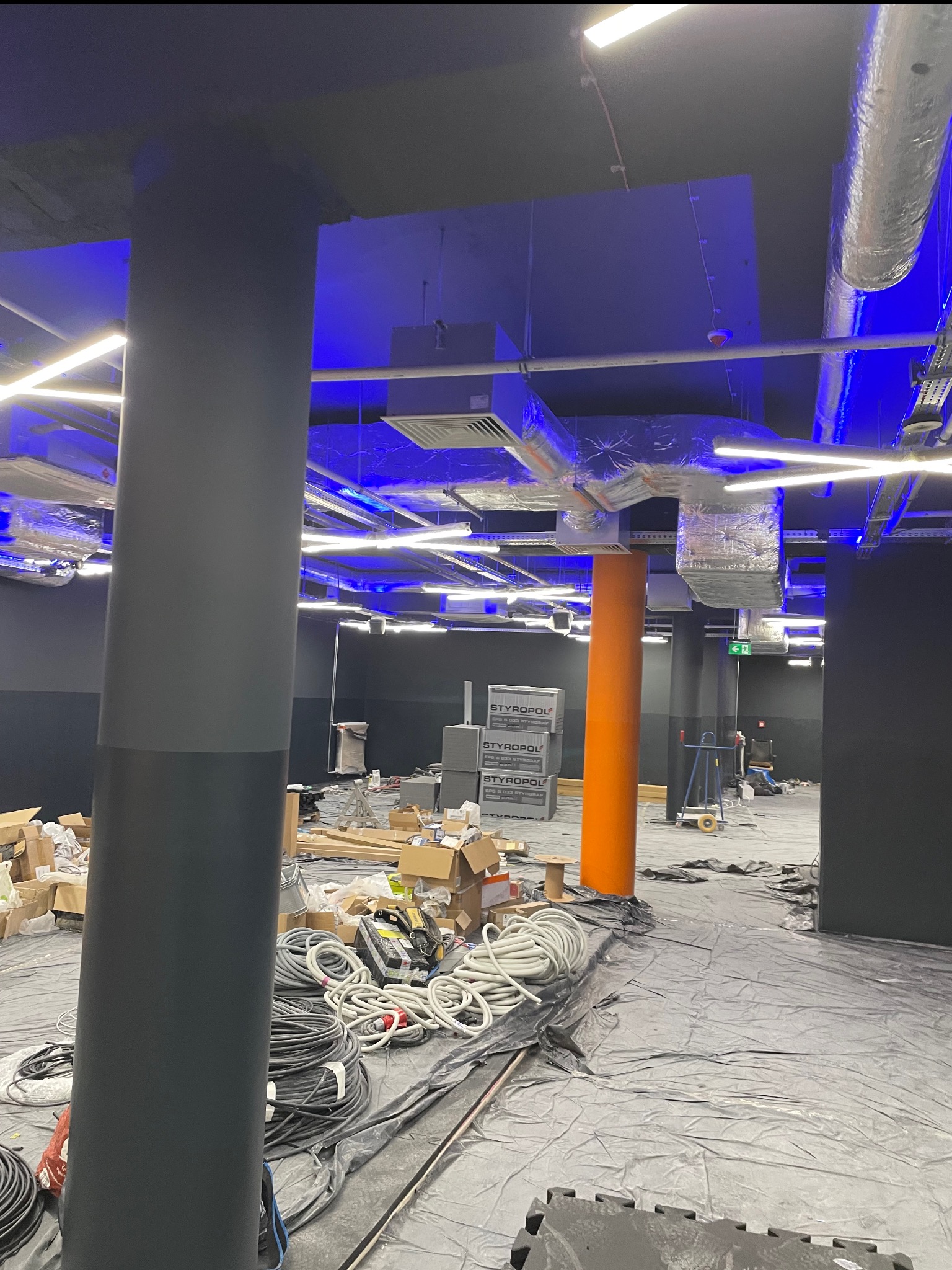 Wnętrze w trakcie prac instalacyjnych: widoczne kable, rury wentylacyjne pod sufitem z ledowym oświetleniem, kolumna w szarym kolorze, folia ochronna na podłodze.
