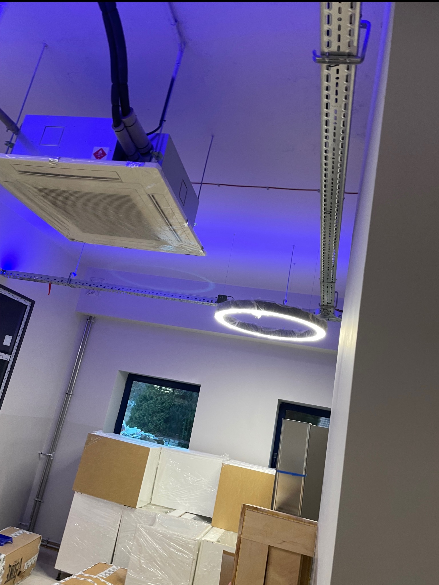 Kasetonowa klimatyzacja sufitowa w folii ochronnej, okrągła lampa LED. Widoczne kable i elementy montażowe. Niebieskie oświetlenie sufitowe.