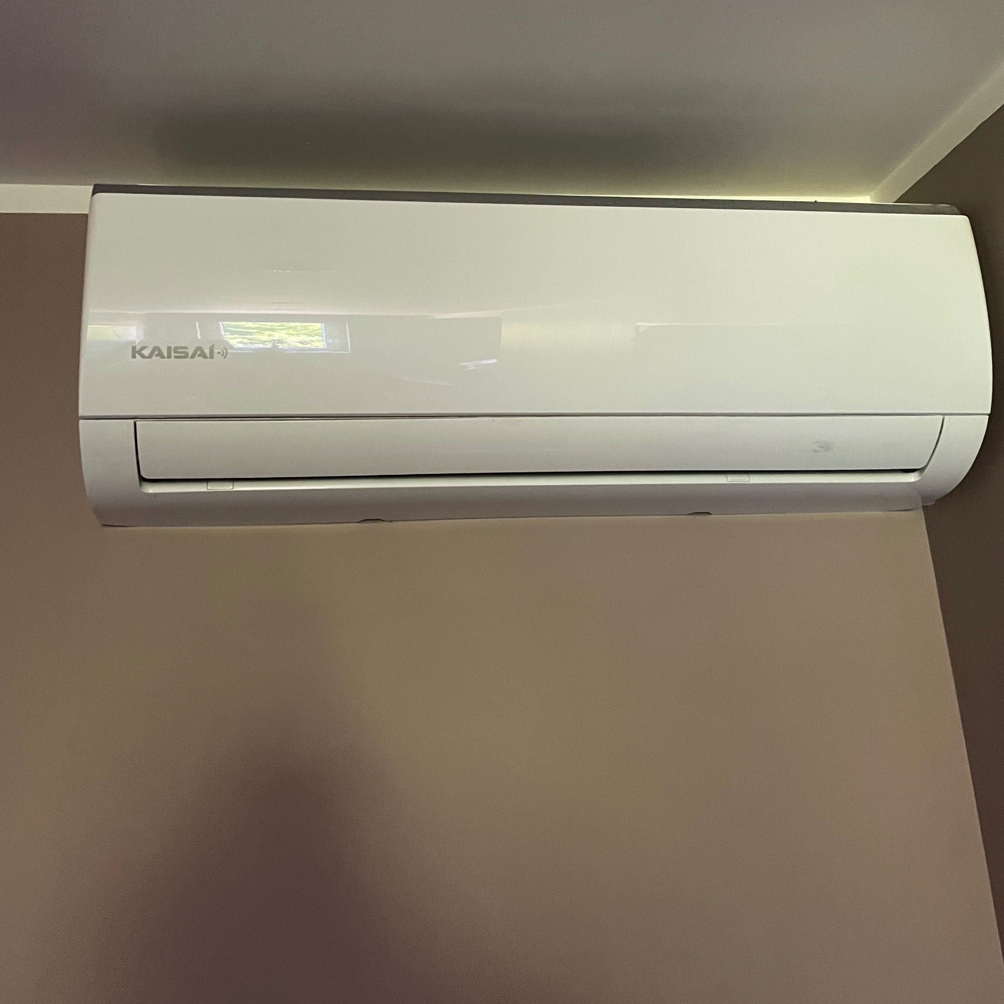 Biały klimatyzator Kaisai na beżowej ścianie, widoczne odbicie okna w obudowie, minimalistyczny design, zamontowany blisko sufitu.