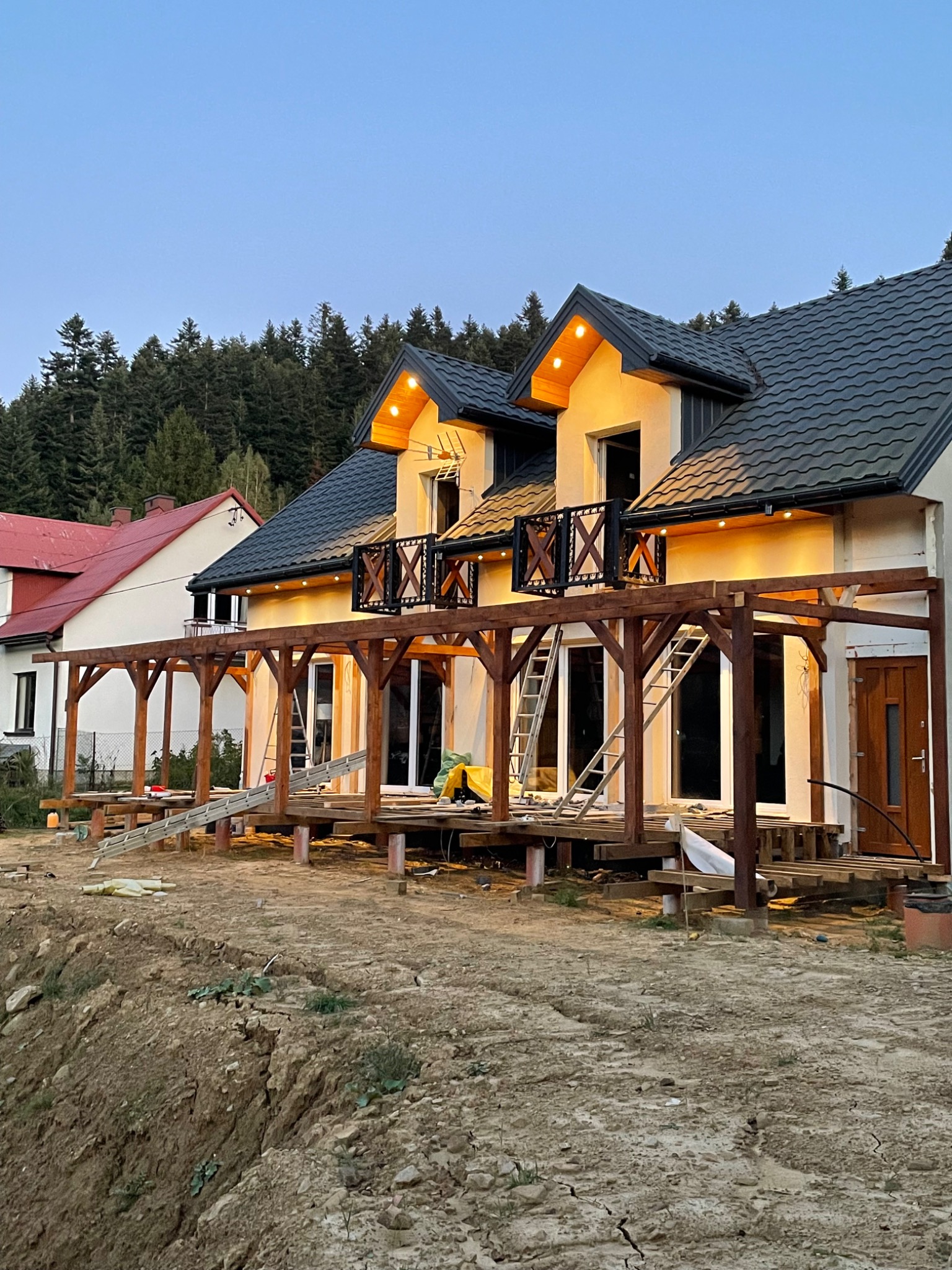 Dwukondygnacyjny dom z drewnianą konstrukcją tarasu i balkonami, oświetlony ciepłym światłem, na tle zalesionego wzgórza, w trakcie budowy.