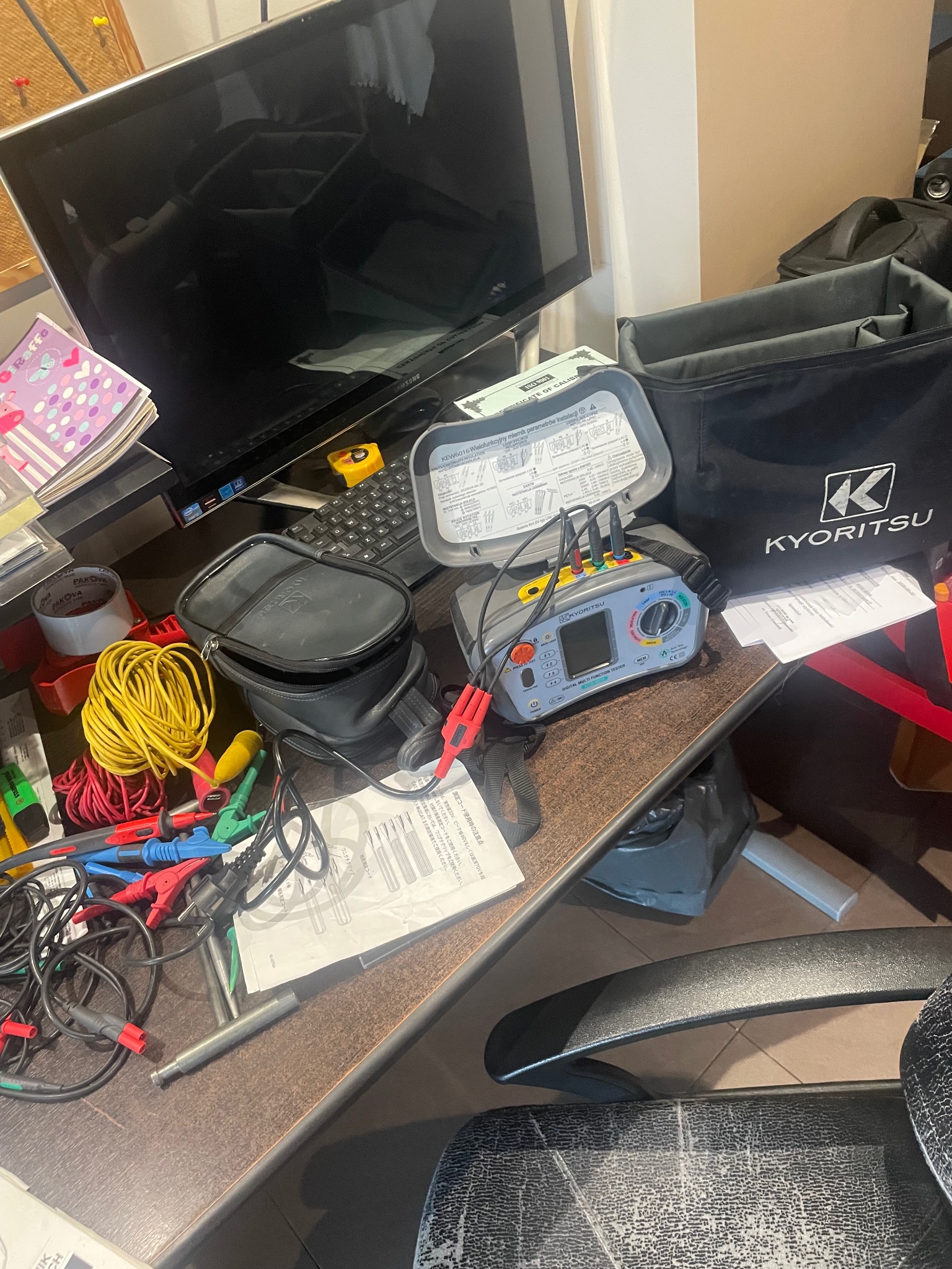 Stanowisko pracy z miernikiem wielofunkcyjnym Kyortisu, przewodami pomiarowymi, dokumentacją i torbą narzędziową na biurku. Widoczne elementy instalacji elektrycznej.