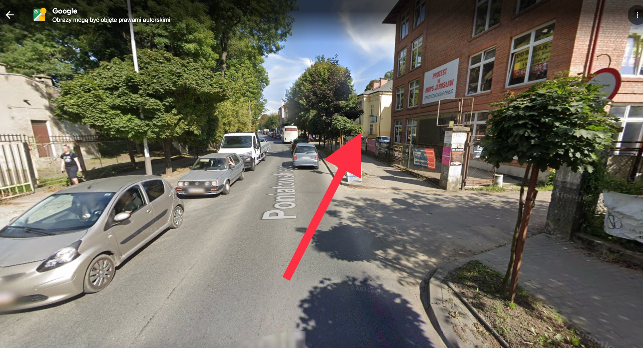 Ulica z budynkiem MOPS Jarosław w tle. Na budynku widoczny baner protestacyjny. Ulica Poniatowskiego z zaparkowanymi samochodami. Słoneczny dzień.