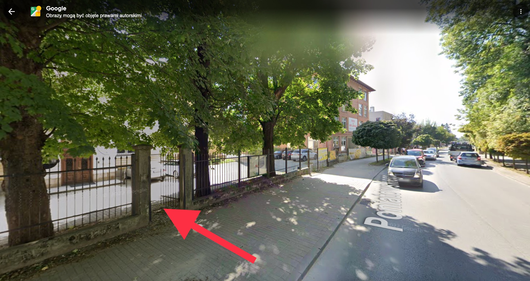 Ulica z chodnikiem i budynkiem w tle, widok z poziomu ulicy, otoczona drzewami i ogrodzeniem. Dzień słoneczny, perspektywa uliczna.