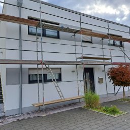 Tomasz Trojanowicz - Biała elewacja domu w trakcie prac wykończeniowych, z widocznym rusztowaniem i zabezpieczeniem podłoża. Fragment schodów zewnętrznych i roślinność w tle.