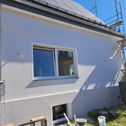 Tomasz Trojanowicz - Boczny widok domu z nową, jasną elewacją o fakturze baranka. Widoczne okna, rusztowania oraz akcesoria malarskie na trawie. Słoneczny dzień.