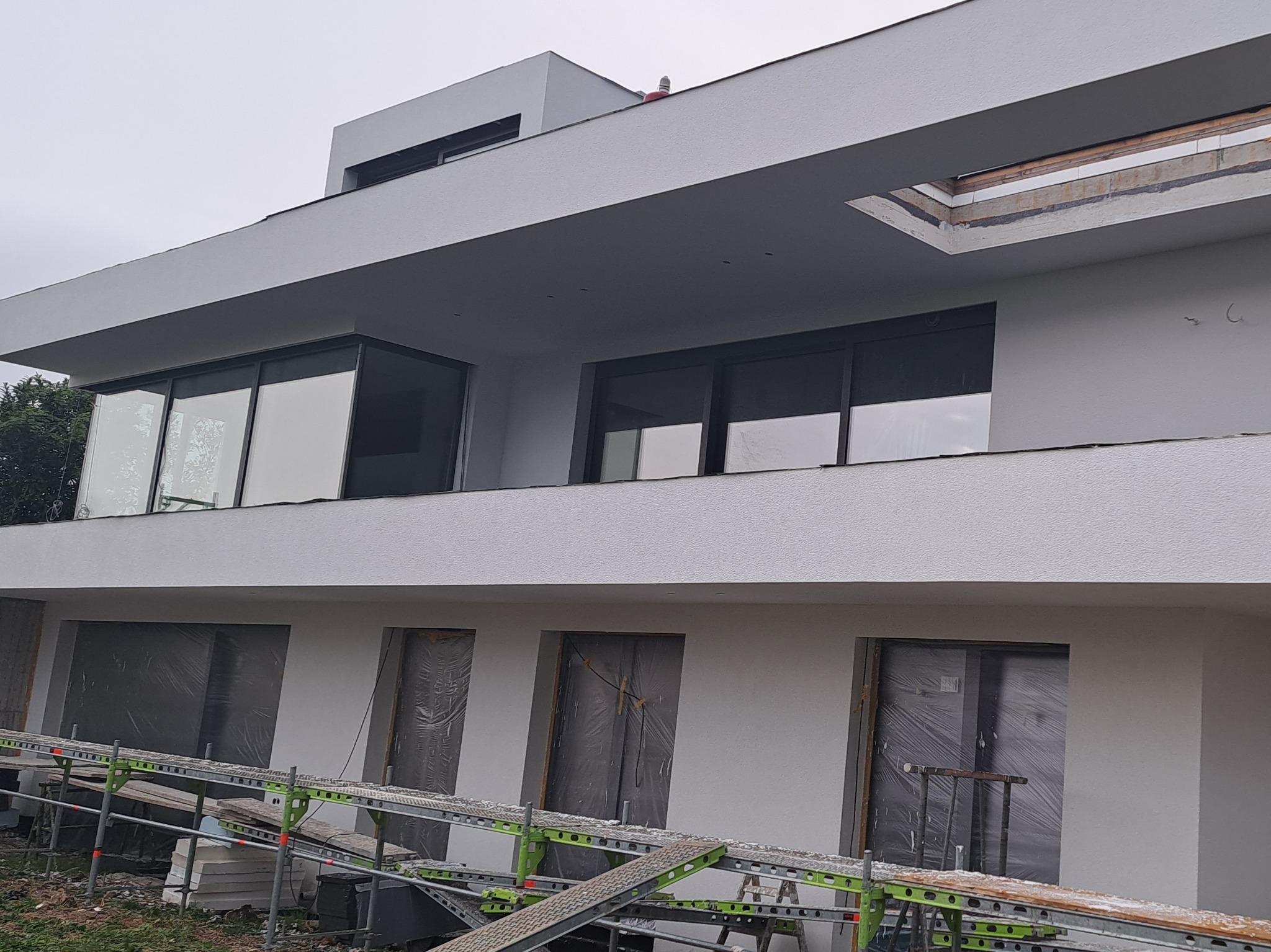 Nowoczesny, dwukondygnacyjny dom po tynkowaniu elewacji na szaro, z rusztowaniem u dołu i oknami zabezpieczonymi folią. Widoczny fragment dachu i błękitne niebo.