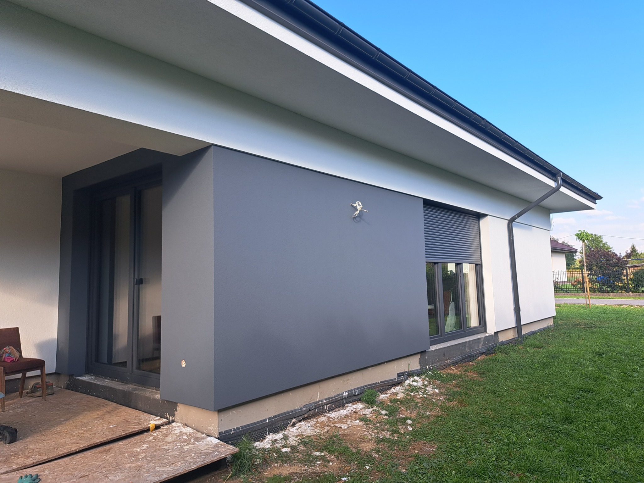 Nowoczesny dom z szarą elewacją, roletami i starannie wykończonymi detalami. Widoczne fragmenty ocieplenia przy gruncie. Ujęcie z zewnątrz, w słoneczny dzień.