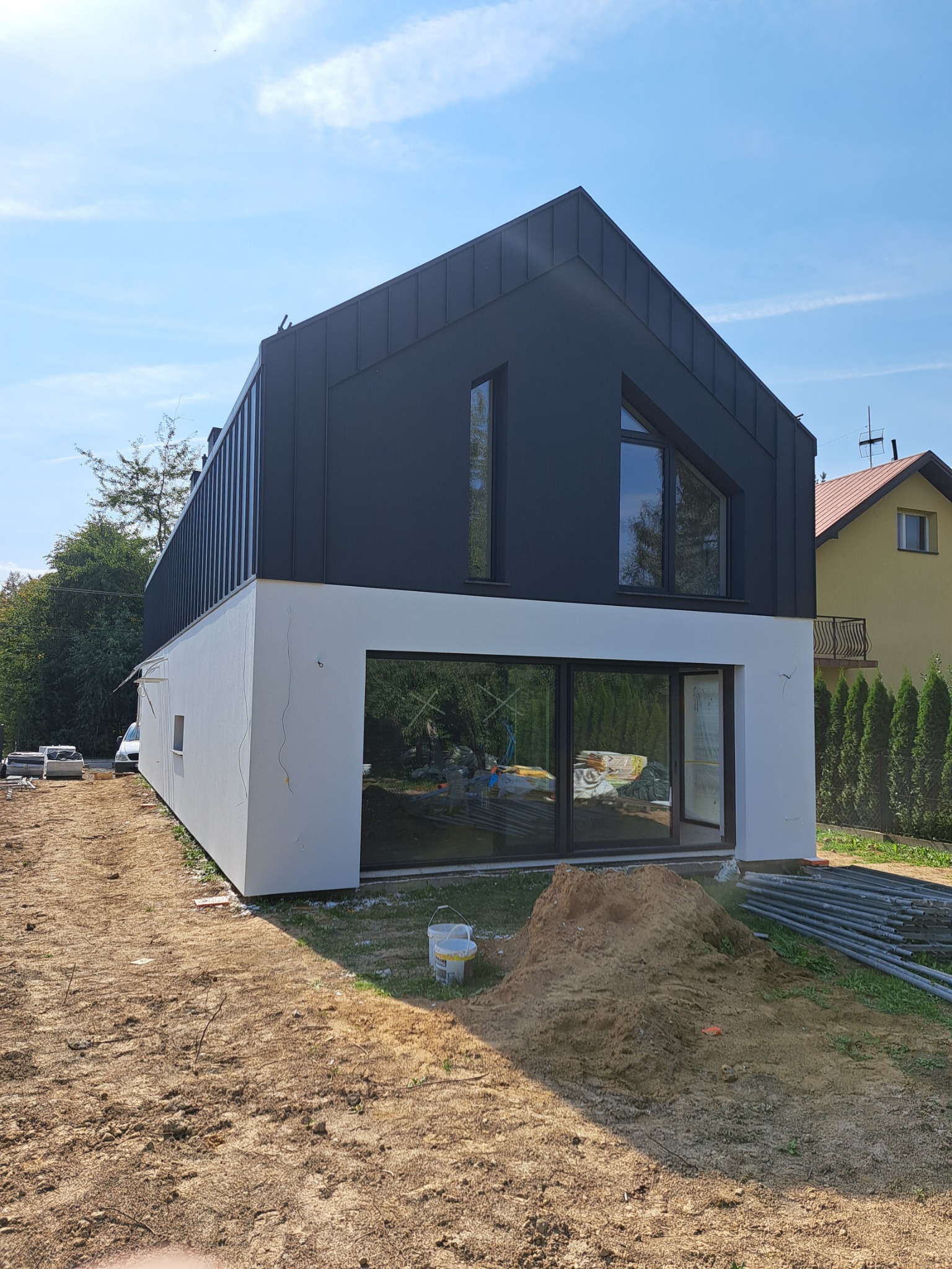 Nowoczesny dom z czarną elewacją i białym parterem w trakcie wykończenia. Widoczne rusztowania i przygotowany teren.