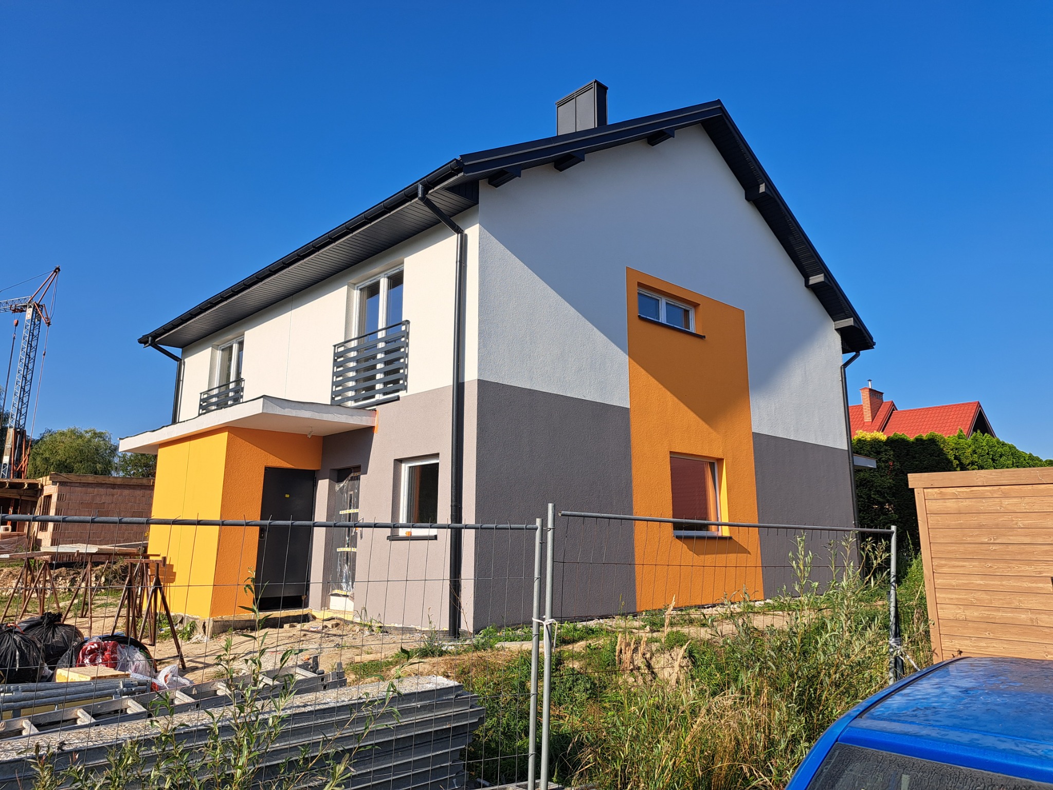 Nowoczesny dom z elewacją w odcieniach szarości, bieli i pomarańczy, widziany z poziomu gruntu. W tle widoczny żuraw budowlany i fragment niebieskiego samochodu.