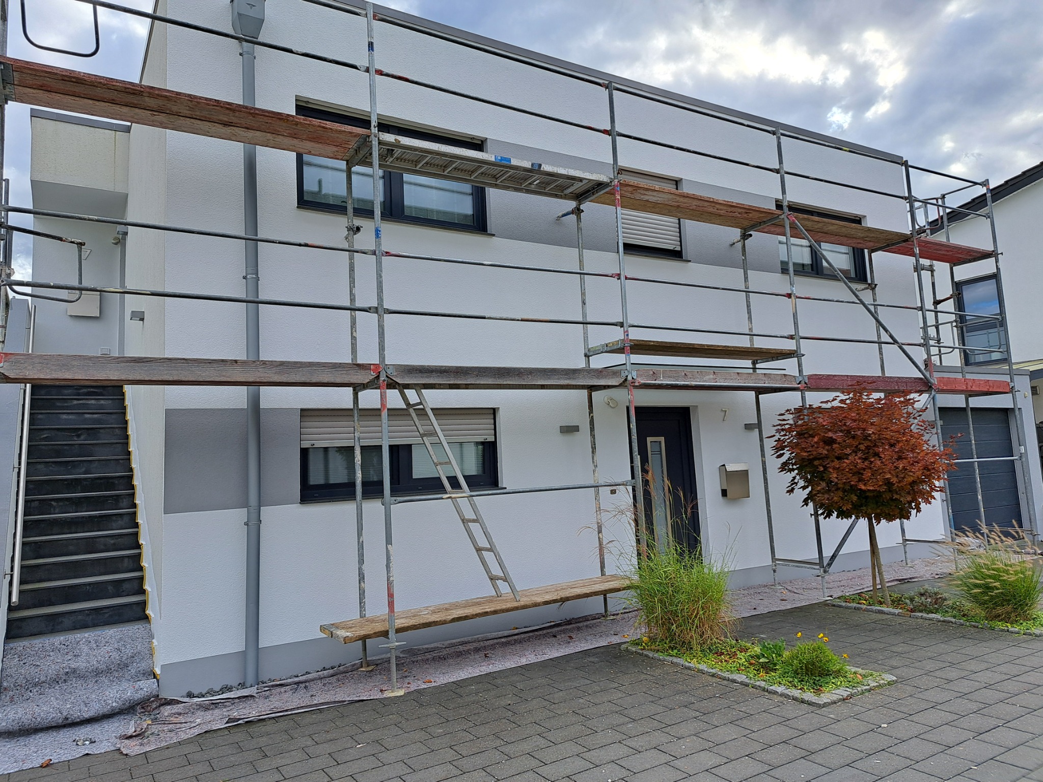 Biała elewacja domu w trakcie prac wykończeniowych, z widocznym rusztowaniem i zabezpieczeniem podłoża. Fragment schodów zewnętrznych i roślinność w tle.