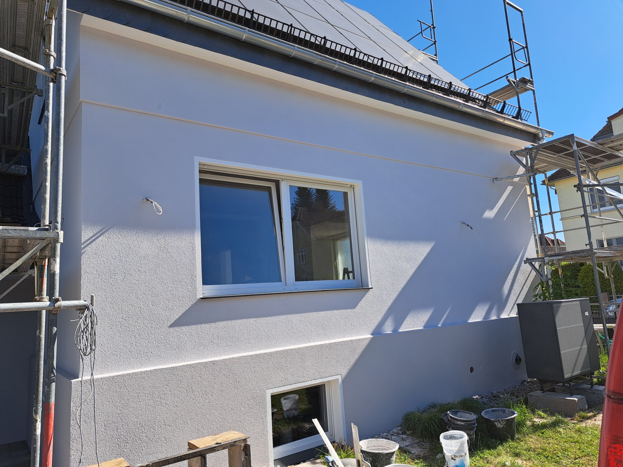 Boczny widok domu z nową, jasną elewacją o fakturze baranka. Widoczne okna, rusztowania oraz akcesoria malarskie na trawie. Słoneczny dzień.