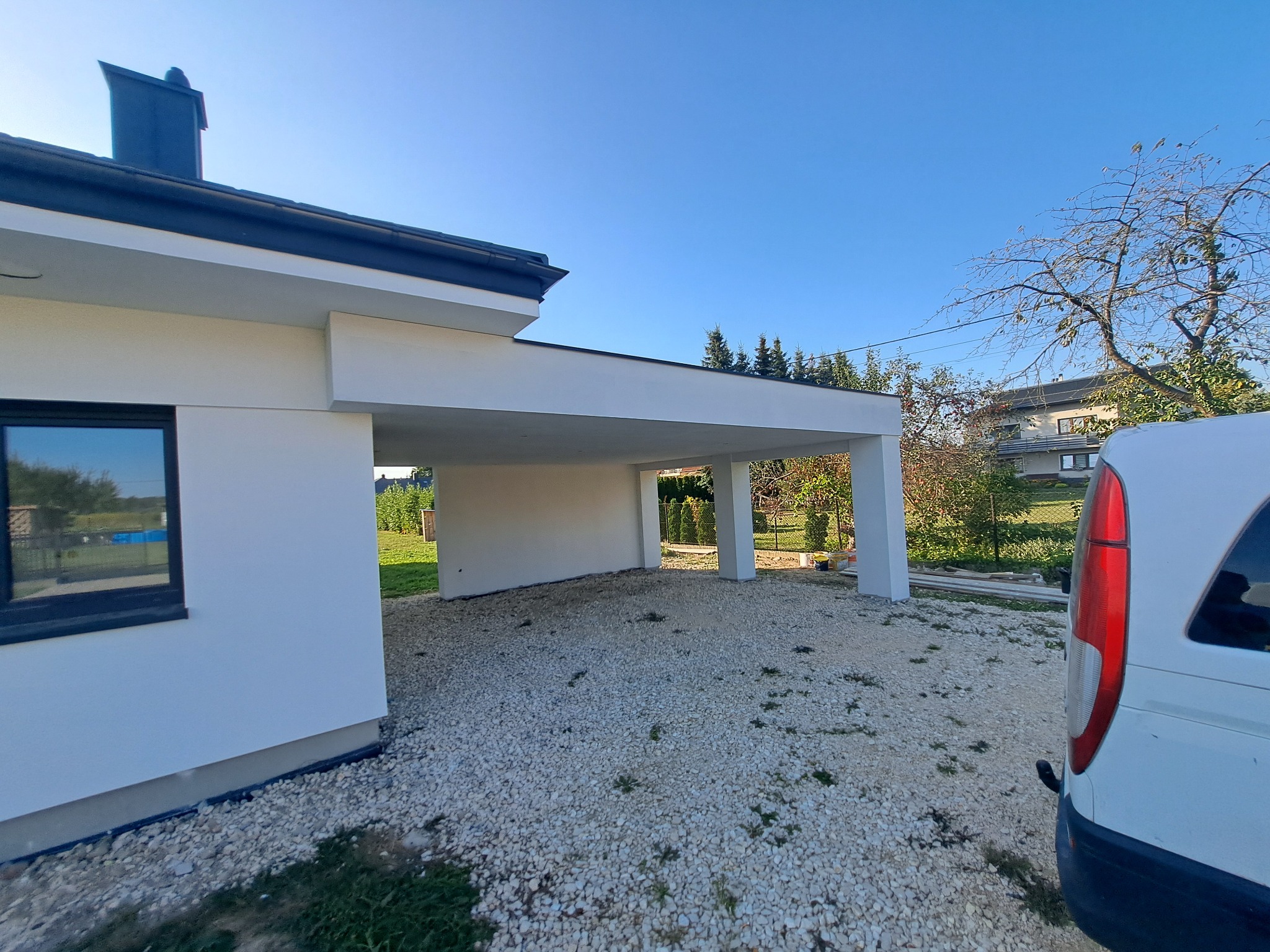 Biały dom z podjazdem wyłożonym żwirem, widok z zewnątrz, fragment samochodu dostawczego po prawej stronie. Elewacja świeżo po malowaniu, w tle zieleń ogrodu.