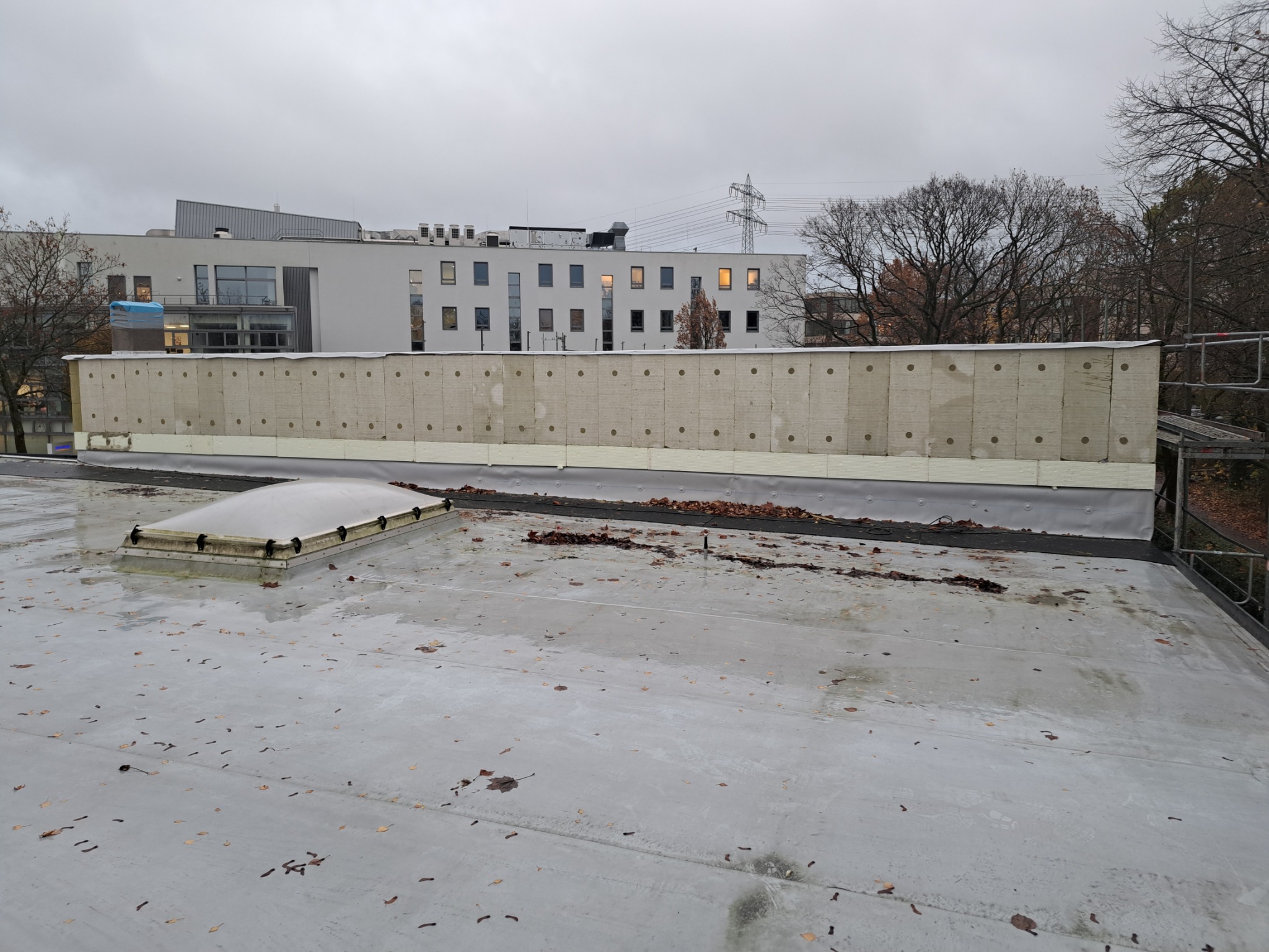 Ocieplenie ściany budynku od strony dachu płaskiego, widoczne płyty izolacyjne z otworami, budynek w tle, pochmurne niebo. Prace na wysokości.