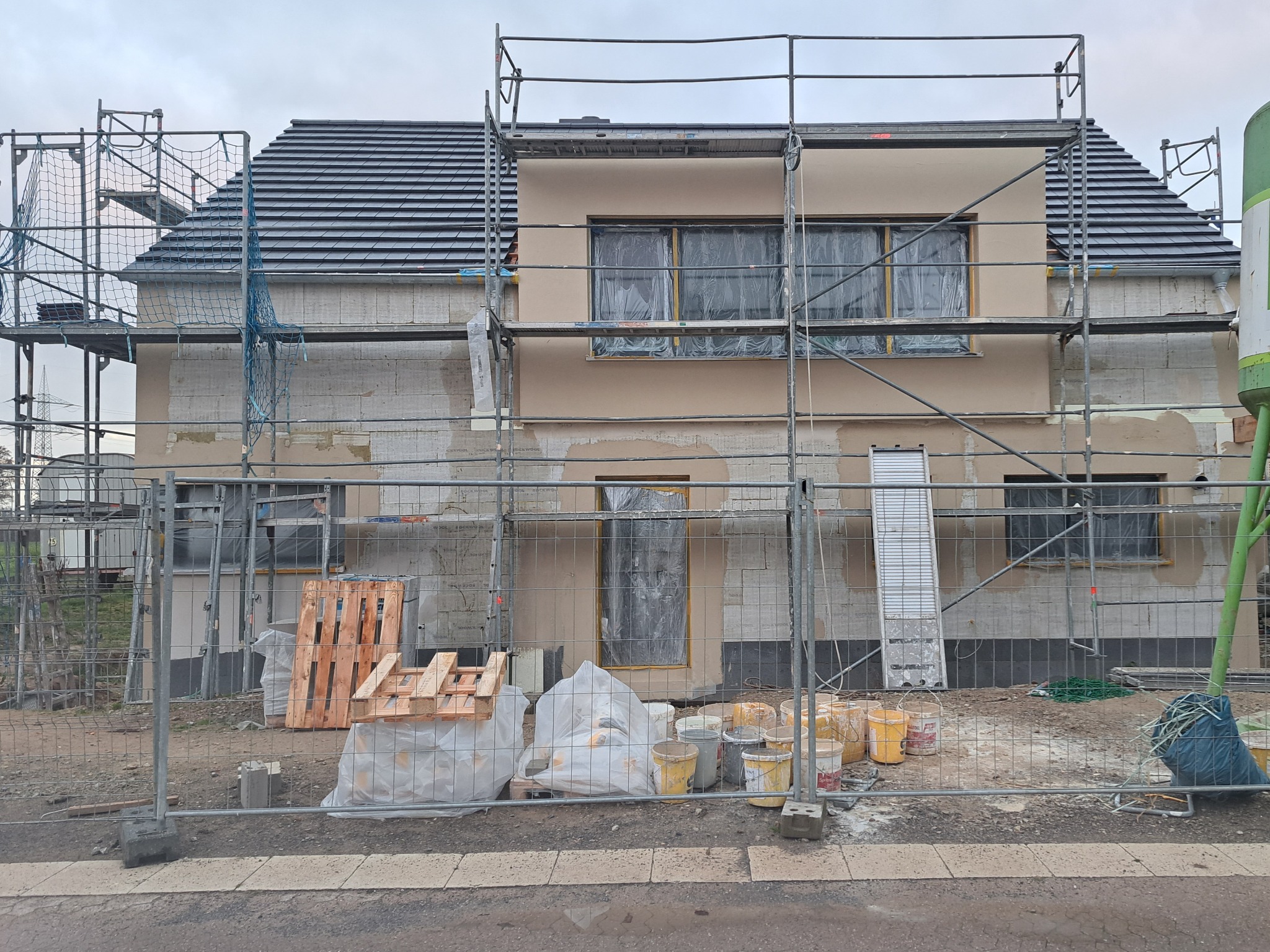 Dom w trakcie ocieplania z rusztowaniem. Część elewacji otynkowana na beżowo, okna zabezpieczone folią. Na dole palety i wiadra z farbą. Widok zza ogrodzenia.