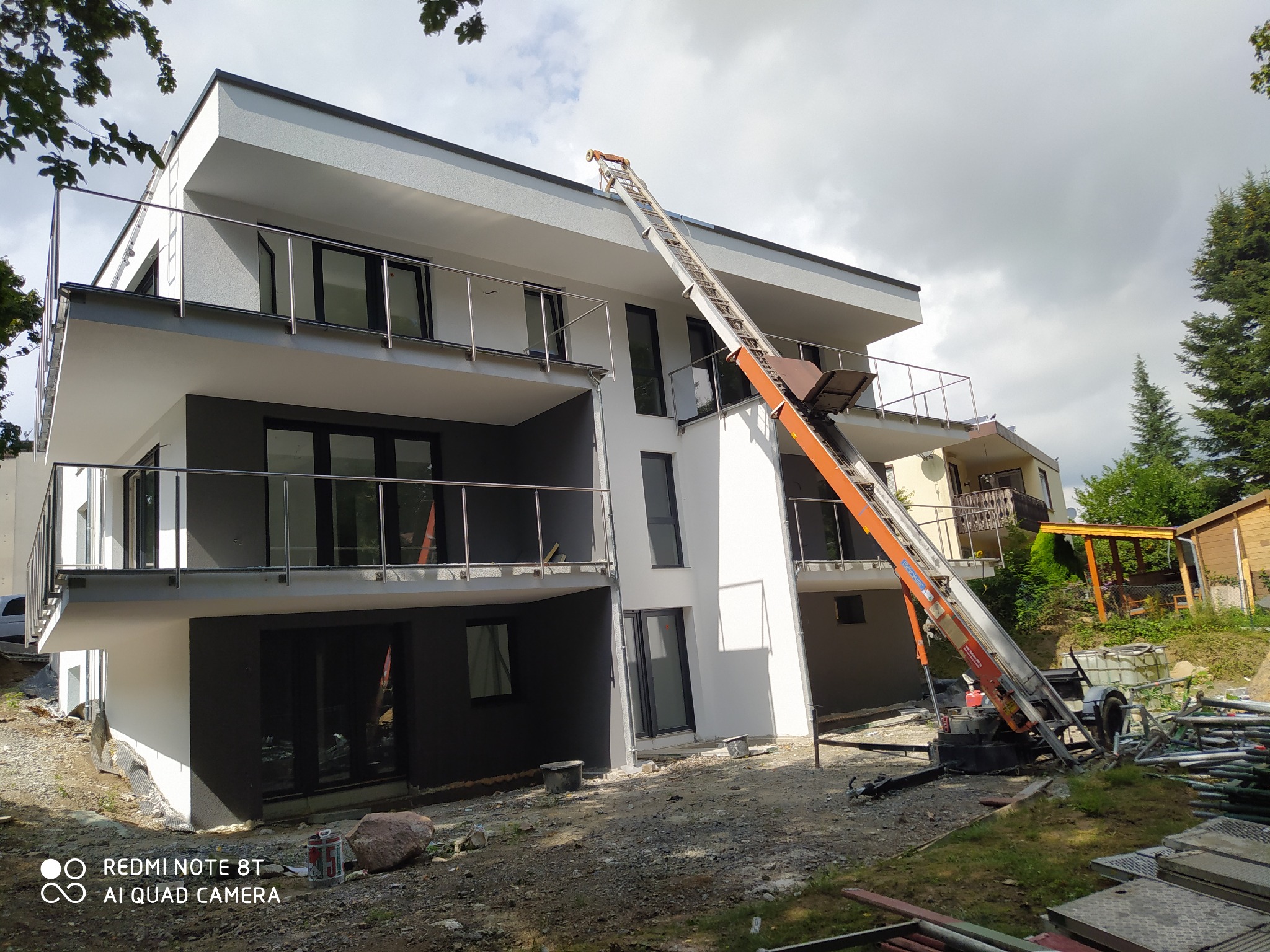 Dwupiętrowy dom z balkonami w trakcie prac wykończeniowych, z podnośnikiem koszowym dostarczającym materiały na dach. Widoczne elementy rusztowania.