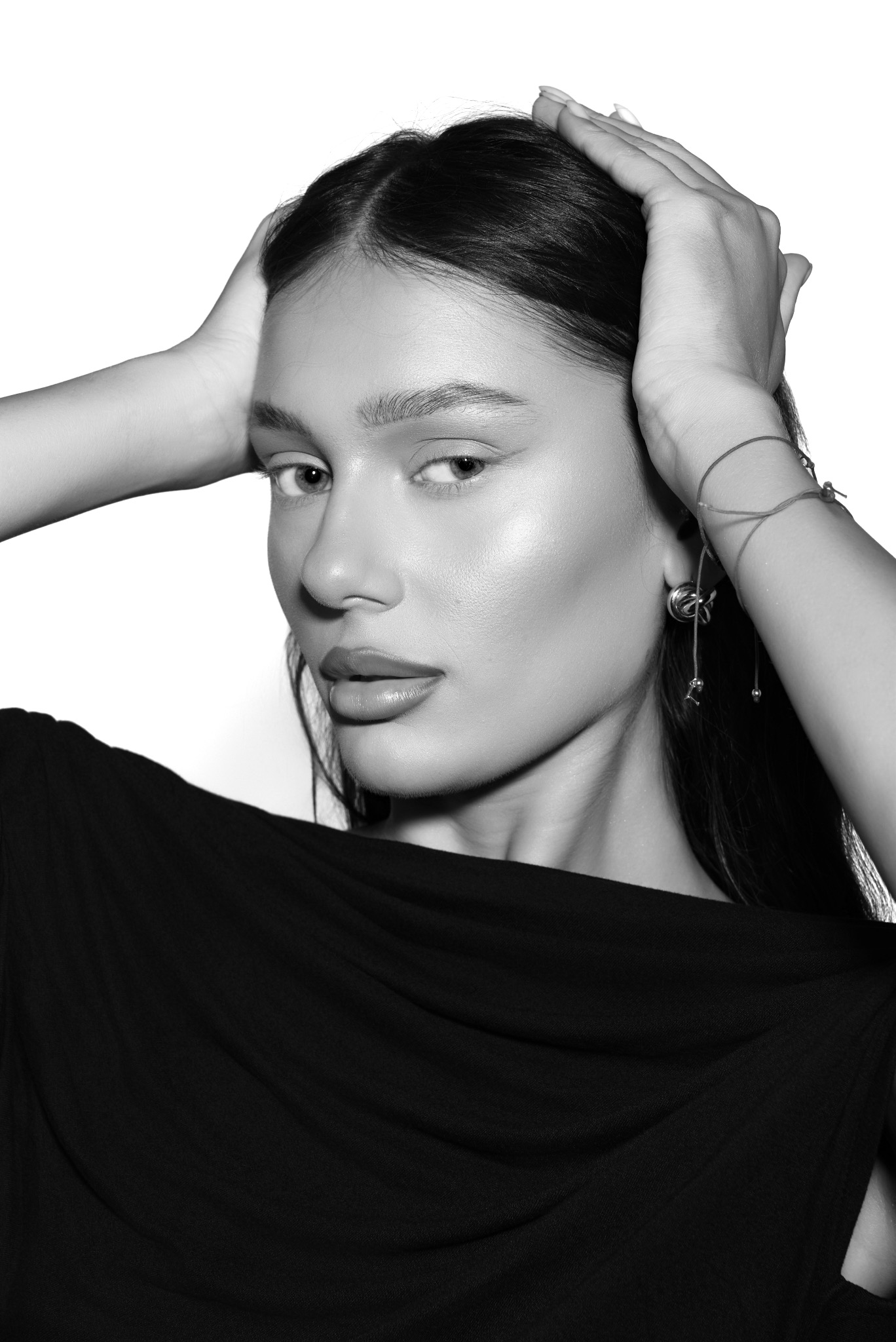 Monochromatyczny portret kobiety z ciemnymi włosami, z rękami uniesionymi do góry, prezentujący makijaż i delikatną biżuterię na białym tle.