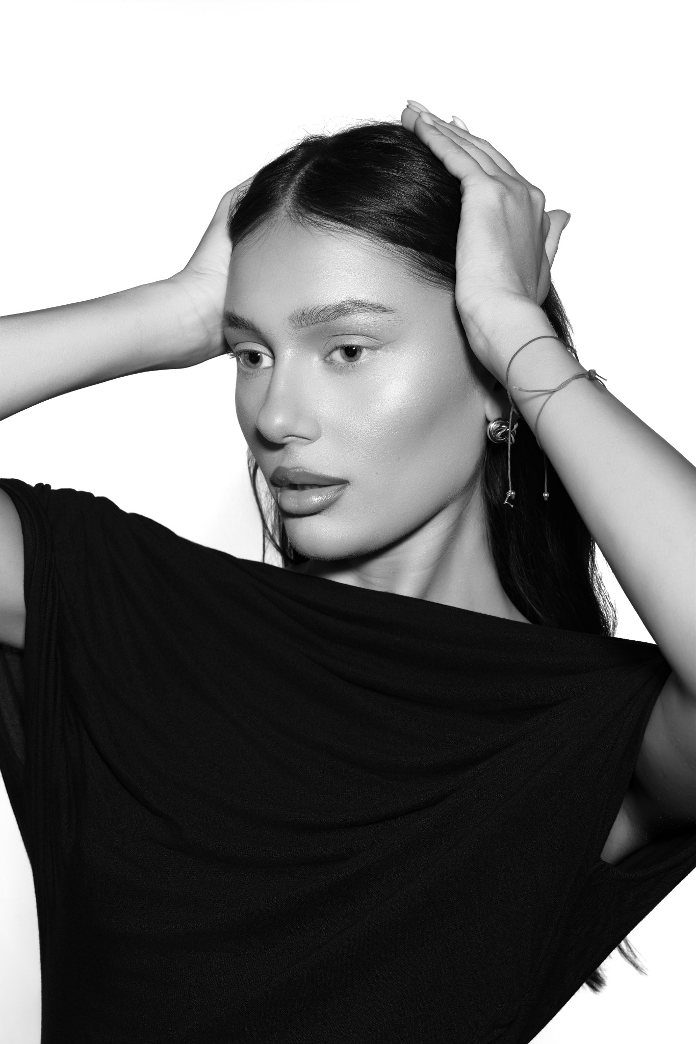 Portret kobiety w czarnej sukni, z rękami uniesionymi do góry, na białym tle. Czarno-biała fotografia podkreśla detale twarzy i biżuterii.