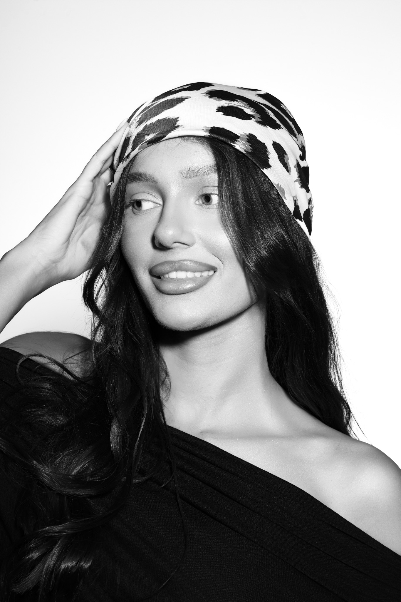 Portret uśmiechniętej kobiety w chuście na głowie, czarno-białe zdjęcie, lekko odwrócona w lewo, z ręką uniesioną do włosów.