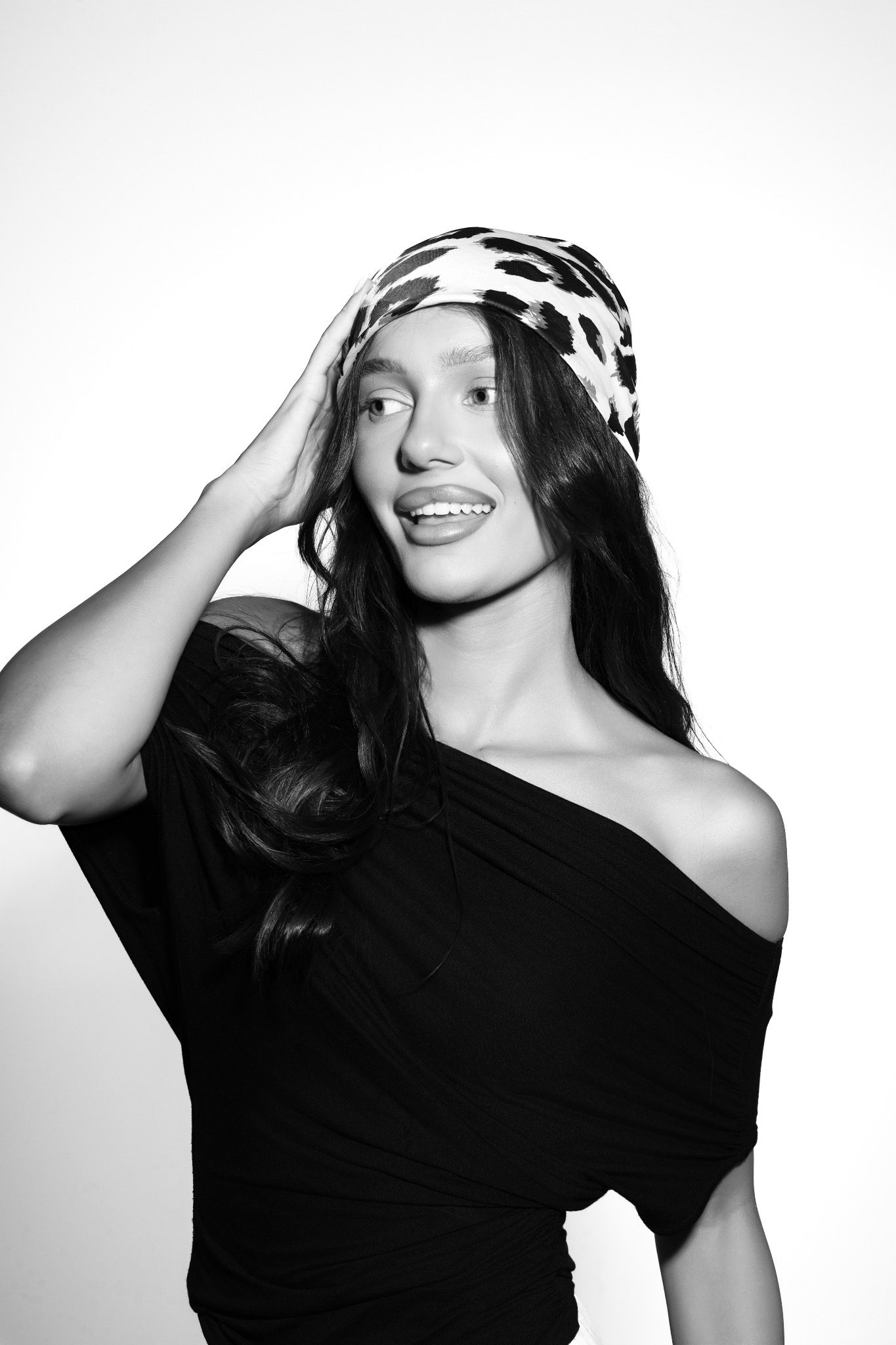 Czarno-białe zdjęcie kobiety z chustą na głowie, ubranej w czarną sukienkę z odkrytym ramieniem, uśmiechniętej i patrzącej w bok, z ręką uniesioną do głowy.