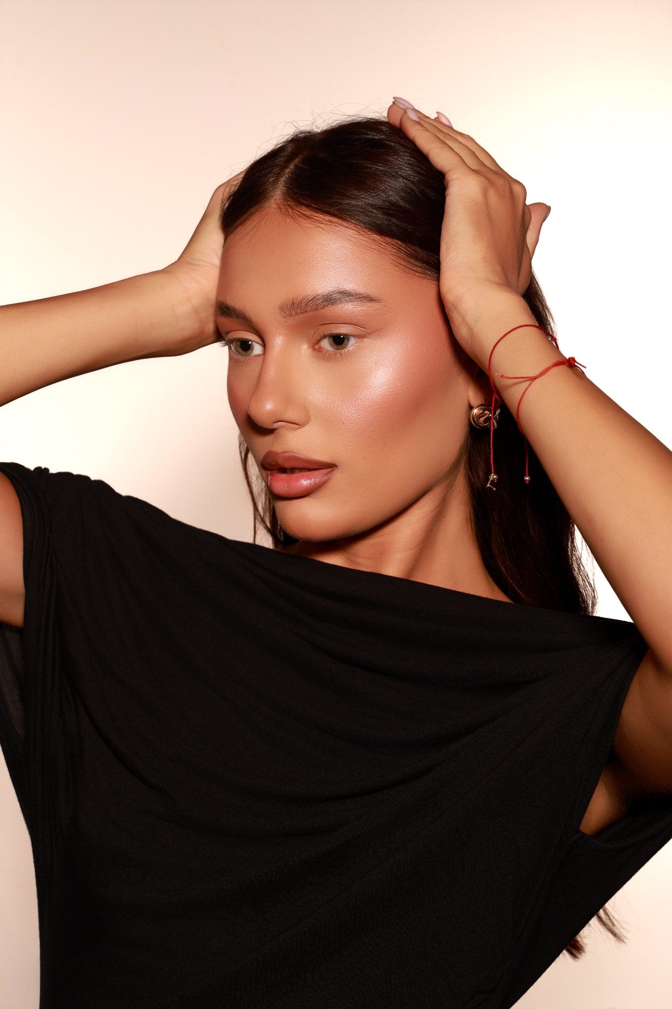 Portret kobiety z ciemnymi włosami, w czarnej sukience, pozującej z rękami uniesionymi do głowy, z delikatną biżuterią i subtelnym makijażem.