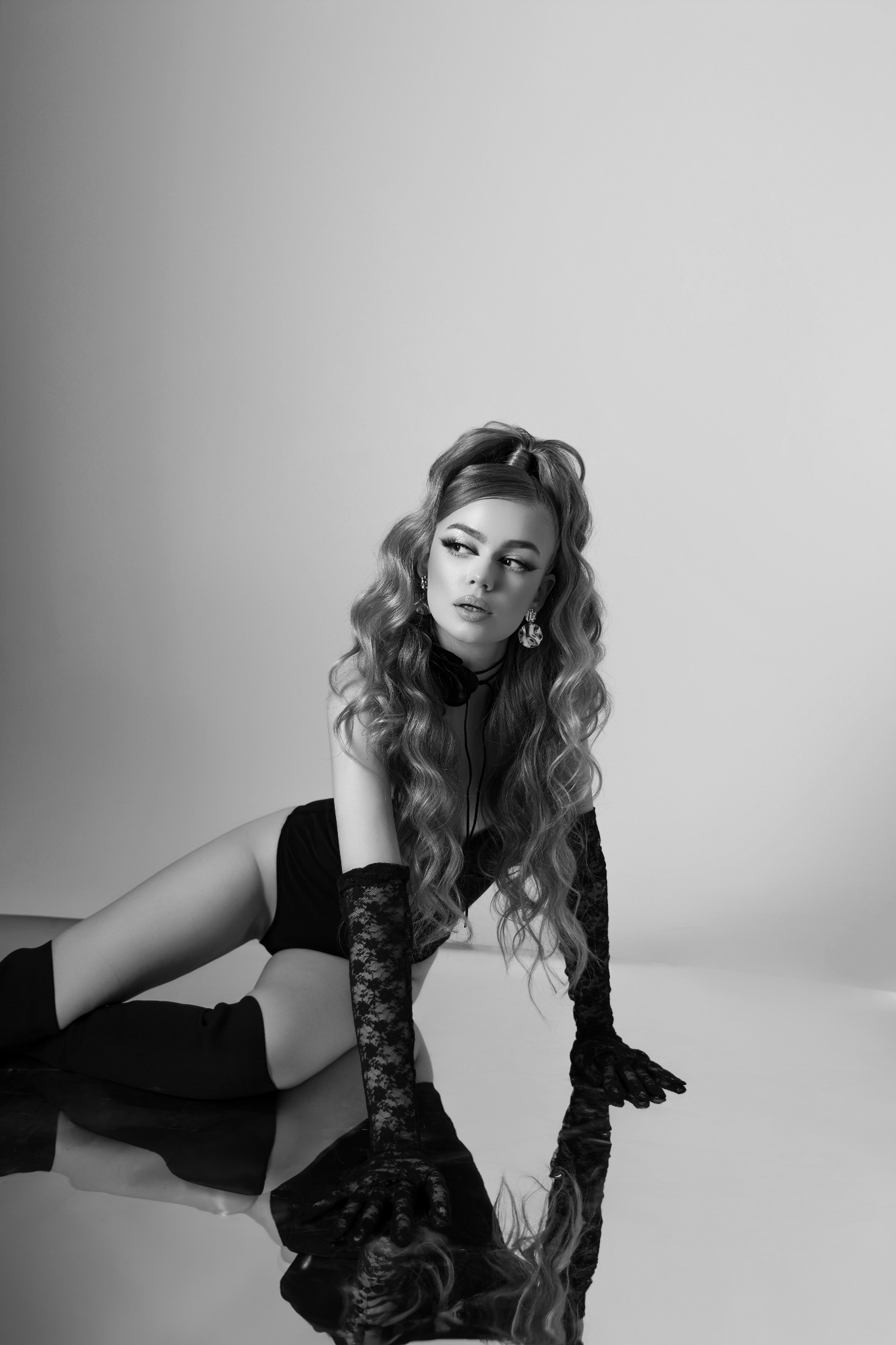 Czarno-biała fotografia kobiety w body, koronkowych rękawiczkach i kozakach, leżącej na lustrzanej powierzchni, z refleksami włosów i stroju, na szarym tle.