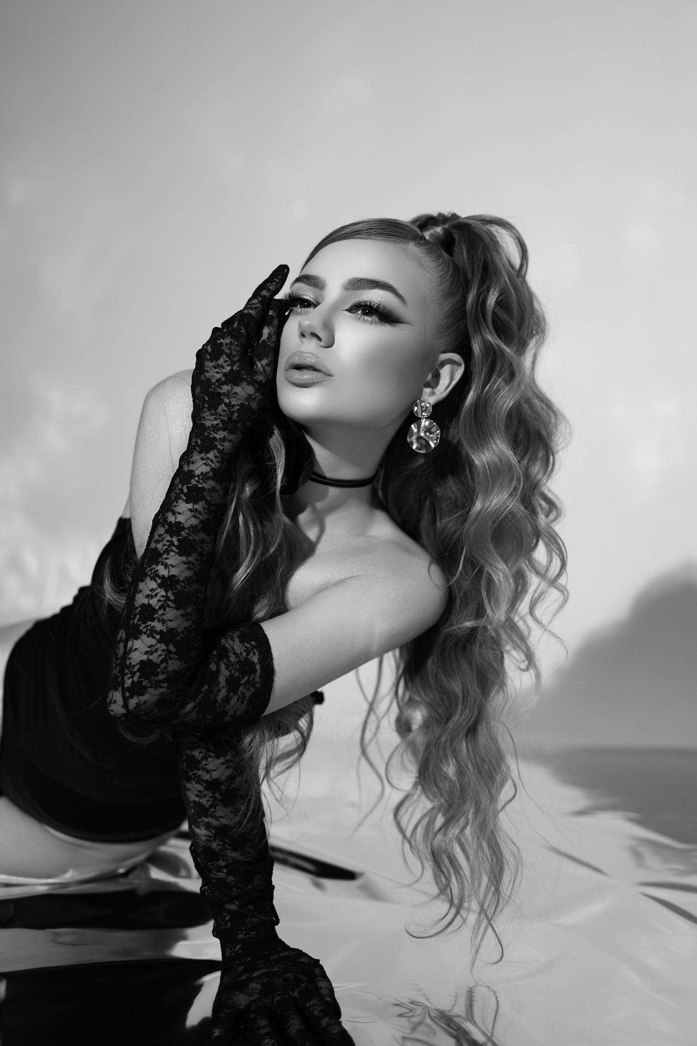 Monochromatyczne ujęcie kobiety w koronkowej rękawiczce, z upiętymi włosami i wyrazistym makijażem, pozującej na błyszczącej powierzchni. Portret w stylu glamour.